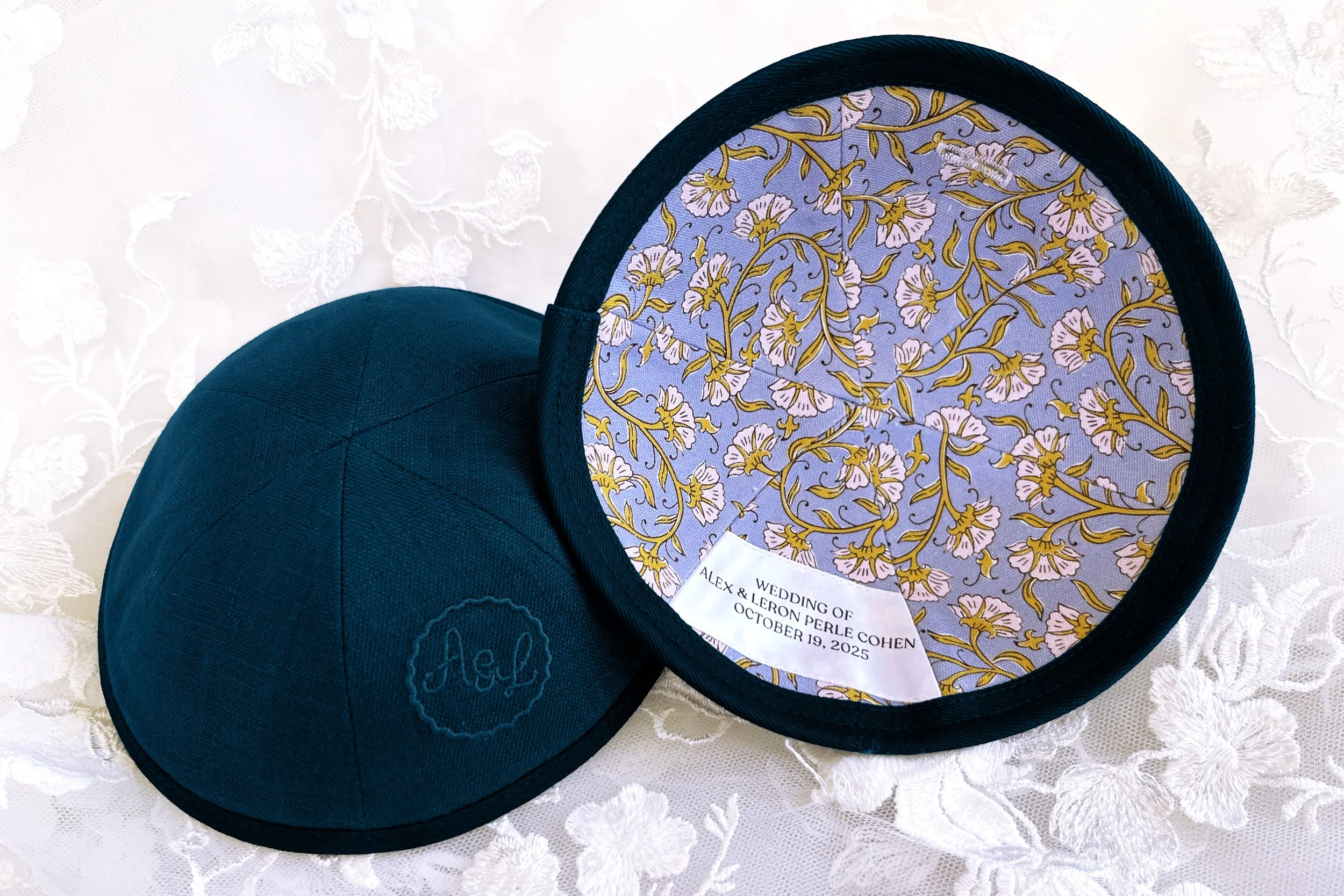 A round dark blue fabric hat with a floral patterned fabric lining and a white tag inside that reads "Wedding of Alex & Leon Perle Cohen October 19, 2025". The hat is on a white lace fabric.