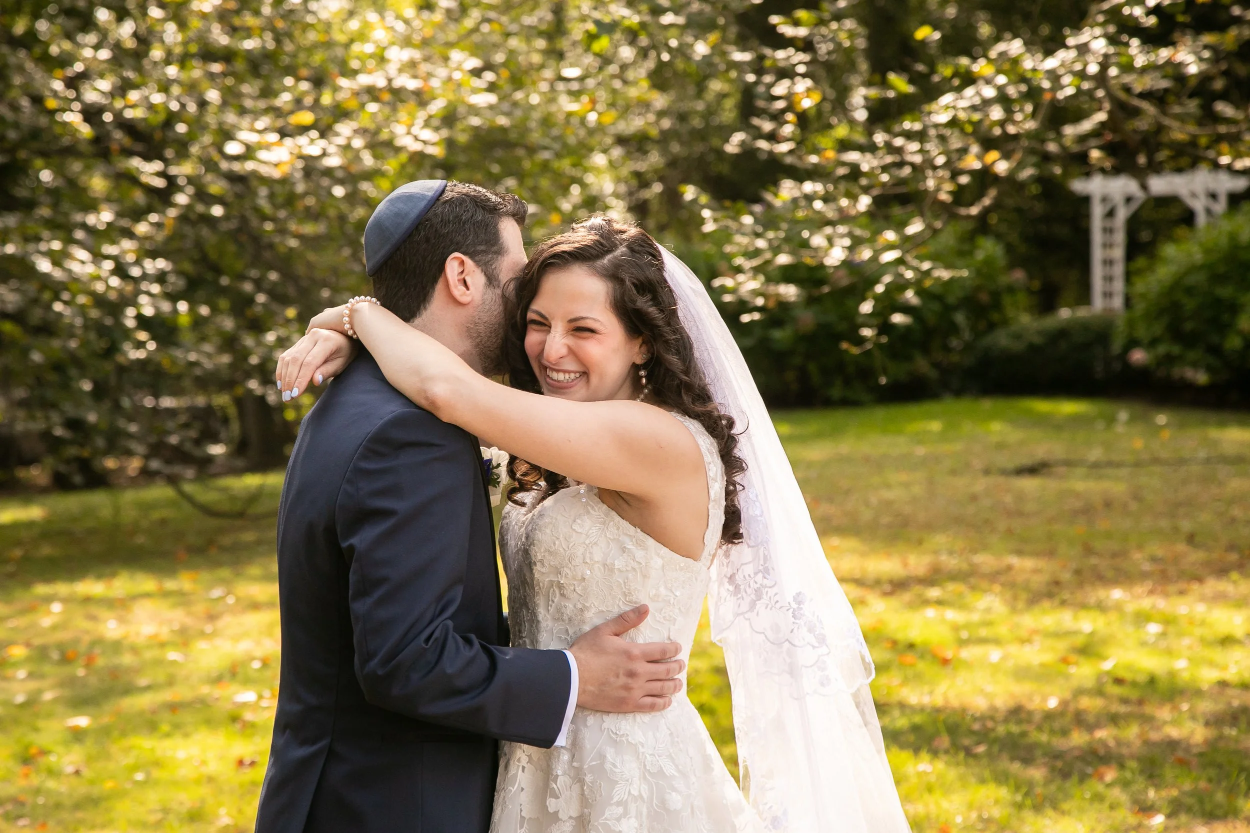 A bride and groom embracing outdoors during a wedding ceremony, with a lush green background and sunlight filtering through trees.