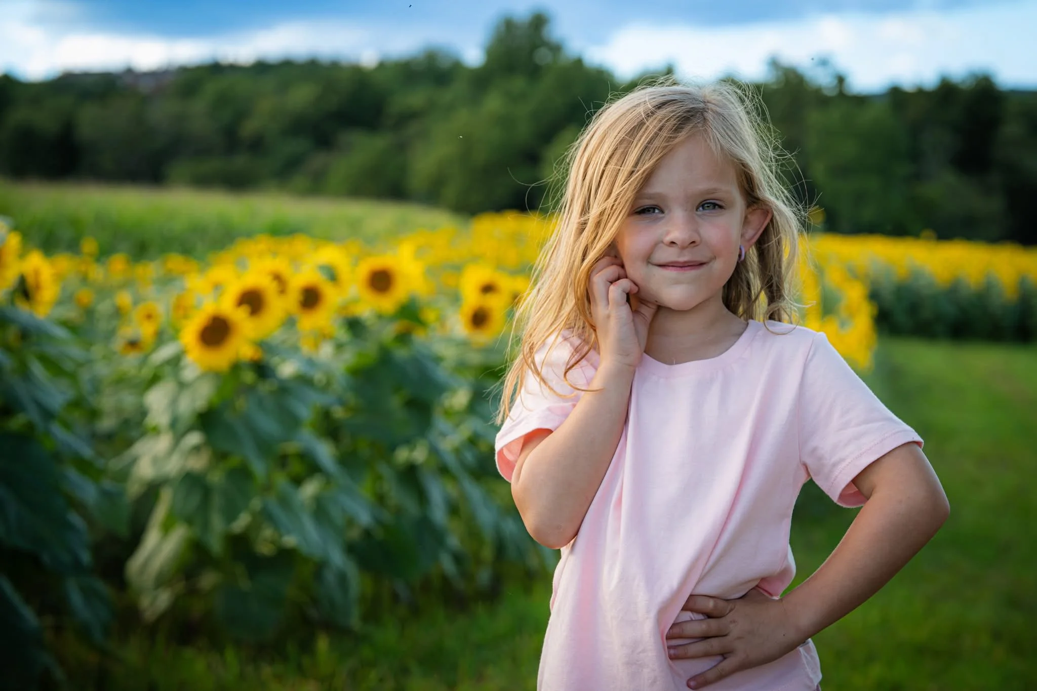 A young girl with blonde hair wearing a pink shirt standing in a field of sunflowers with trees and a cloudy sky in the background.