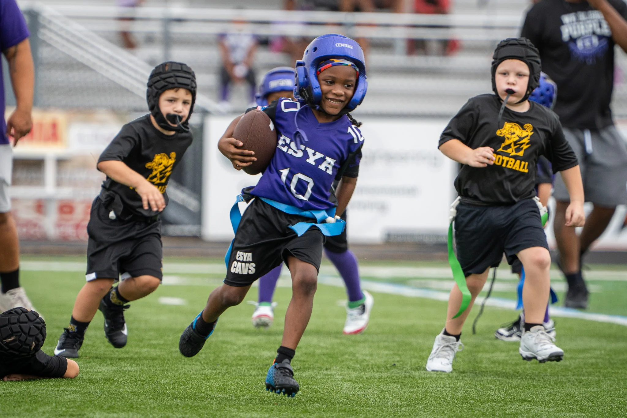 Young children playing tackle football on a field, wearing helmets and athletic gear, with one girl holding a football.