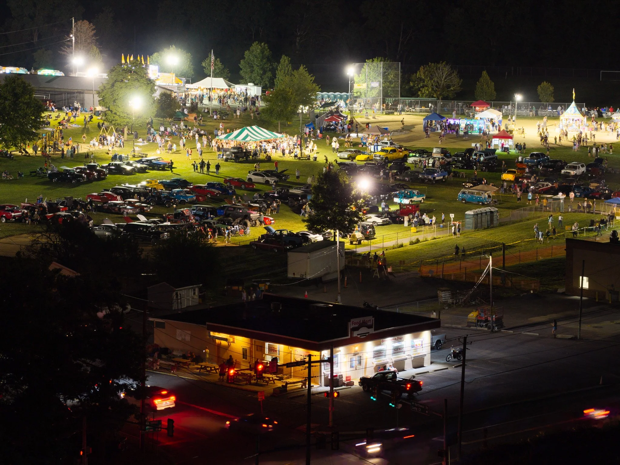 A lively nighttime scene at a fairground with bright lights, tents, and people walking around. There are numerous cars parked and some moving on the street below.