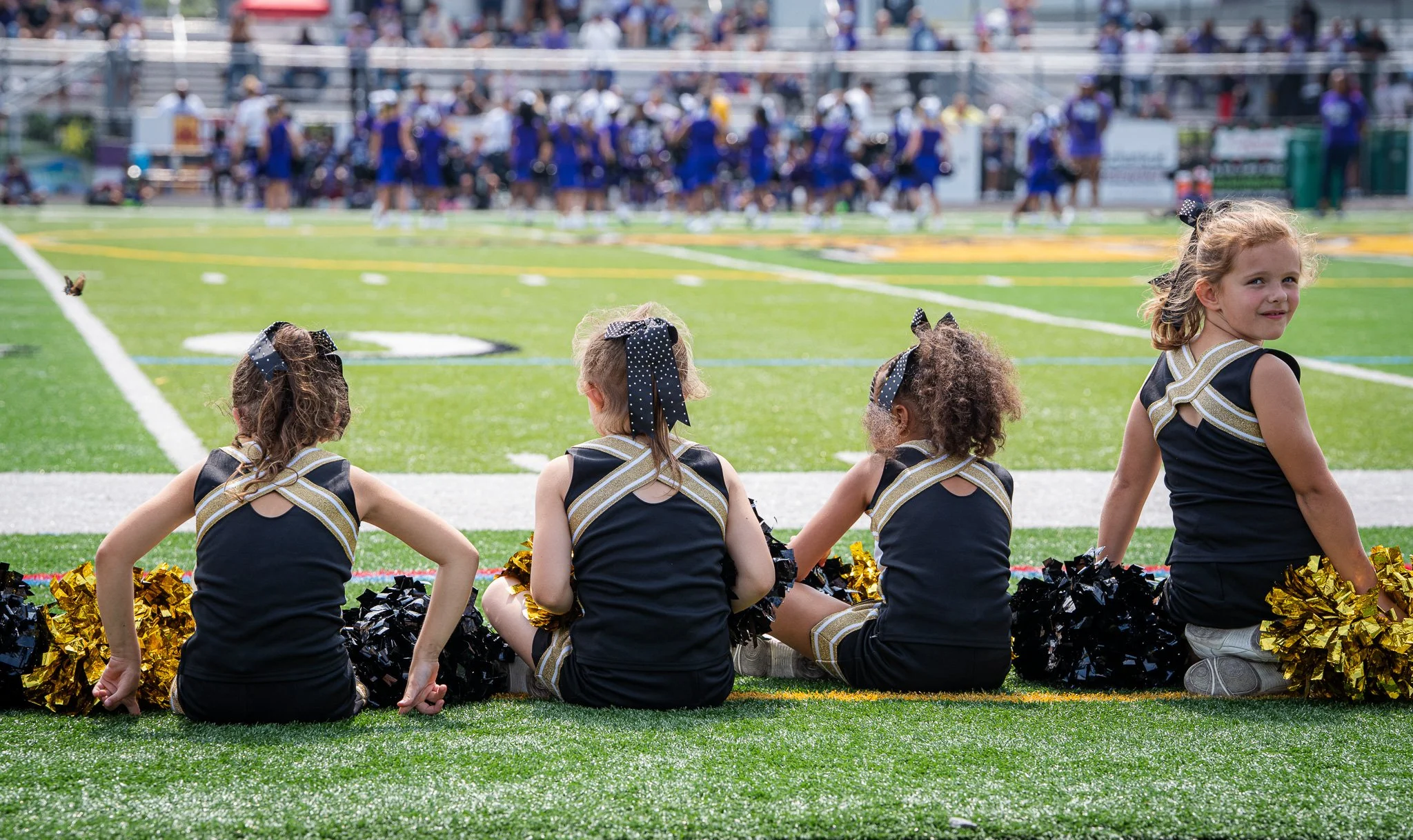 Four young cheerleaders sitting on the sideline of a football field, watching a game. They are dressed in black and gold uniforms with matching bows in their hair, holding pom-poms. In the background, a football team in purple and white uniforms is o