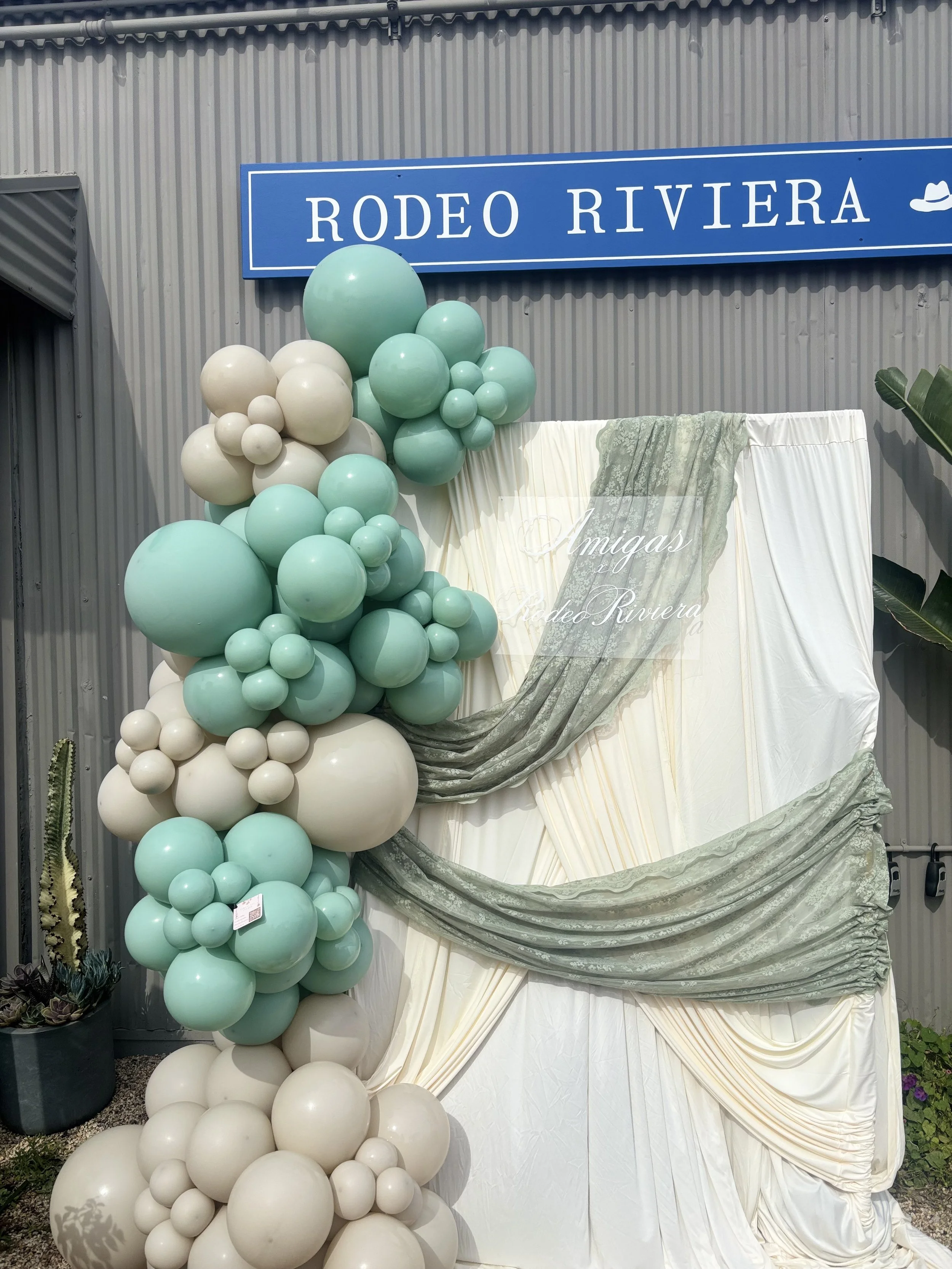 A decorative display with numerous balloons in shades of green and white arranged in a cascading manner. Behind the balloons, there is a white draped fabric with an overlay of green patterned fabric and the text 'Amigas x Rodeo Riviera' written on it