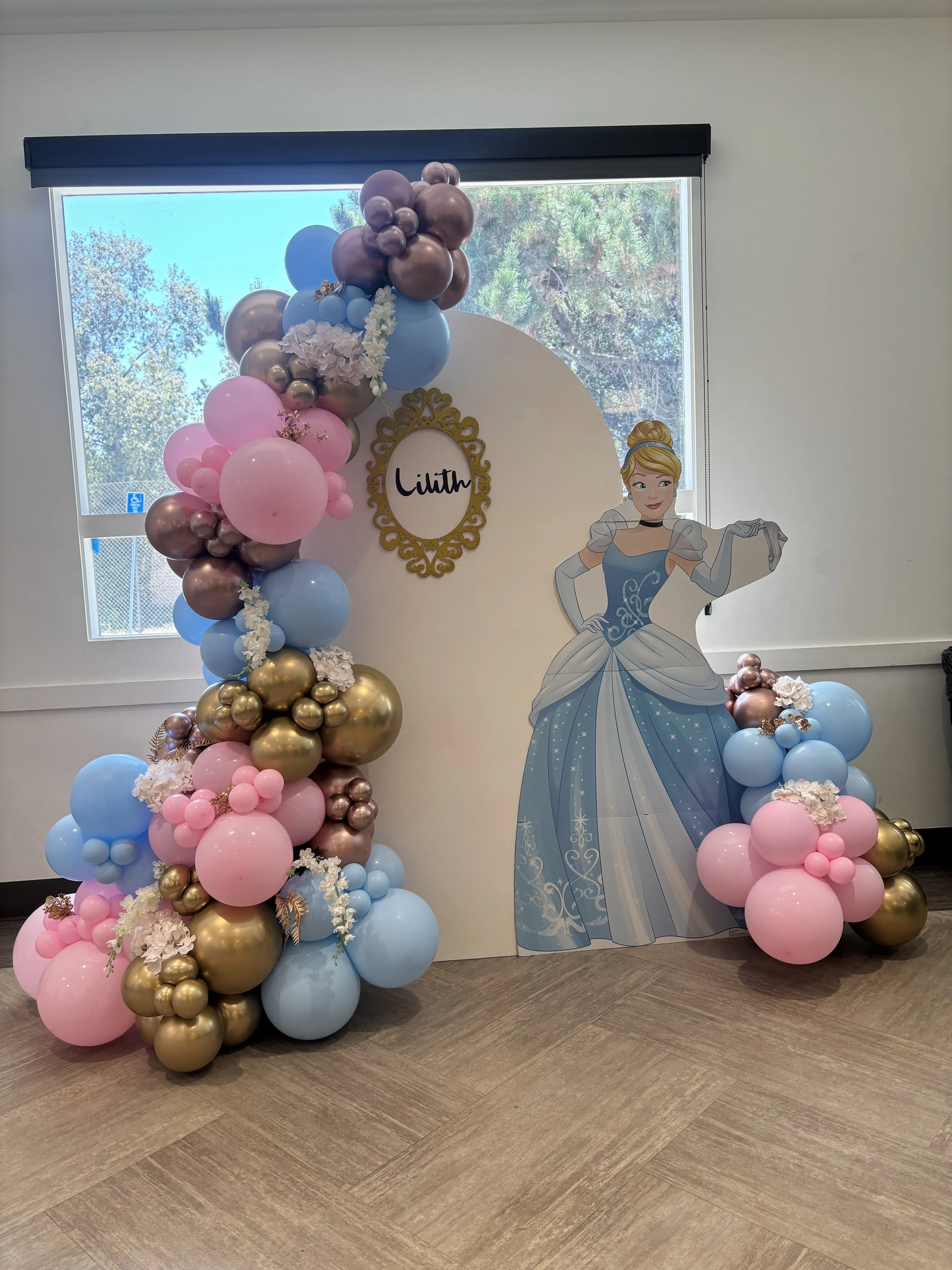Decorative setup with pink, blue, gold, and silver balloons arranged around a white backdrop with a princess illustration of Cinderella, and a gold ornate frame with the name 'Lilith' written inside.