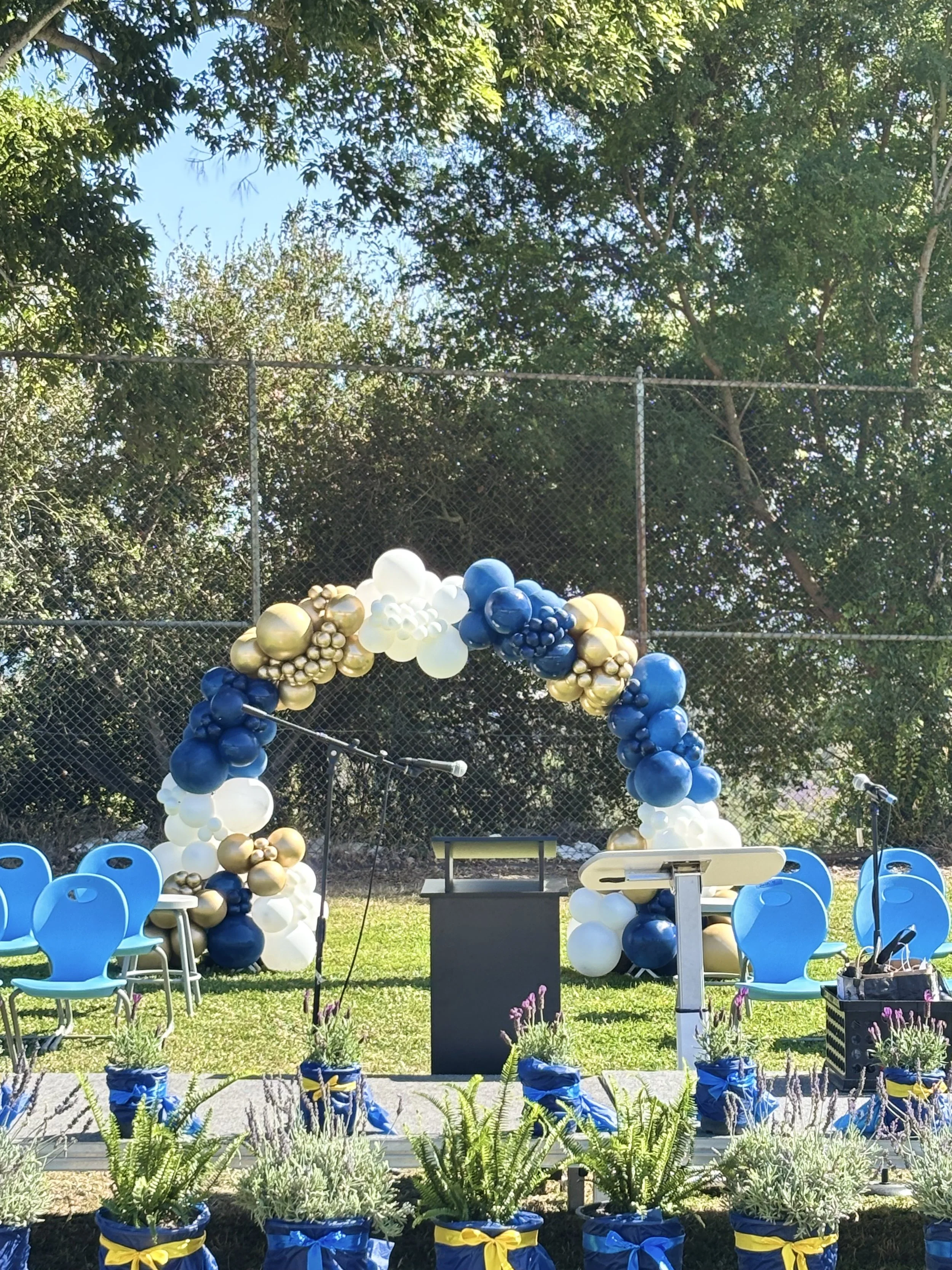 Outdoor memorial setup with balloon arch and chairs, decorated in blue, white, and gold, with potted lavender plants in front and a black podium at the center, surrounded by trees and clear sky.