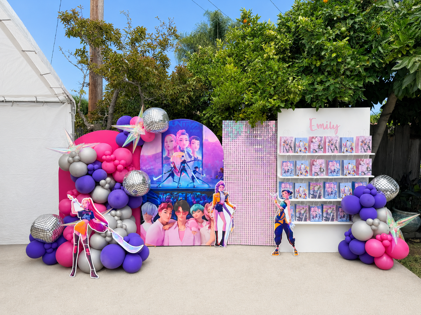 Decorated outdoor space with pink, purple, and silver balloons, photo display of animated characters, and a gift shelf with pink and purple themed bags labeled 'Emily' for a celebration or party.
