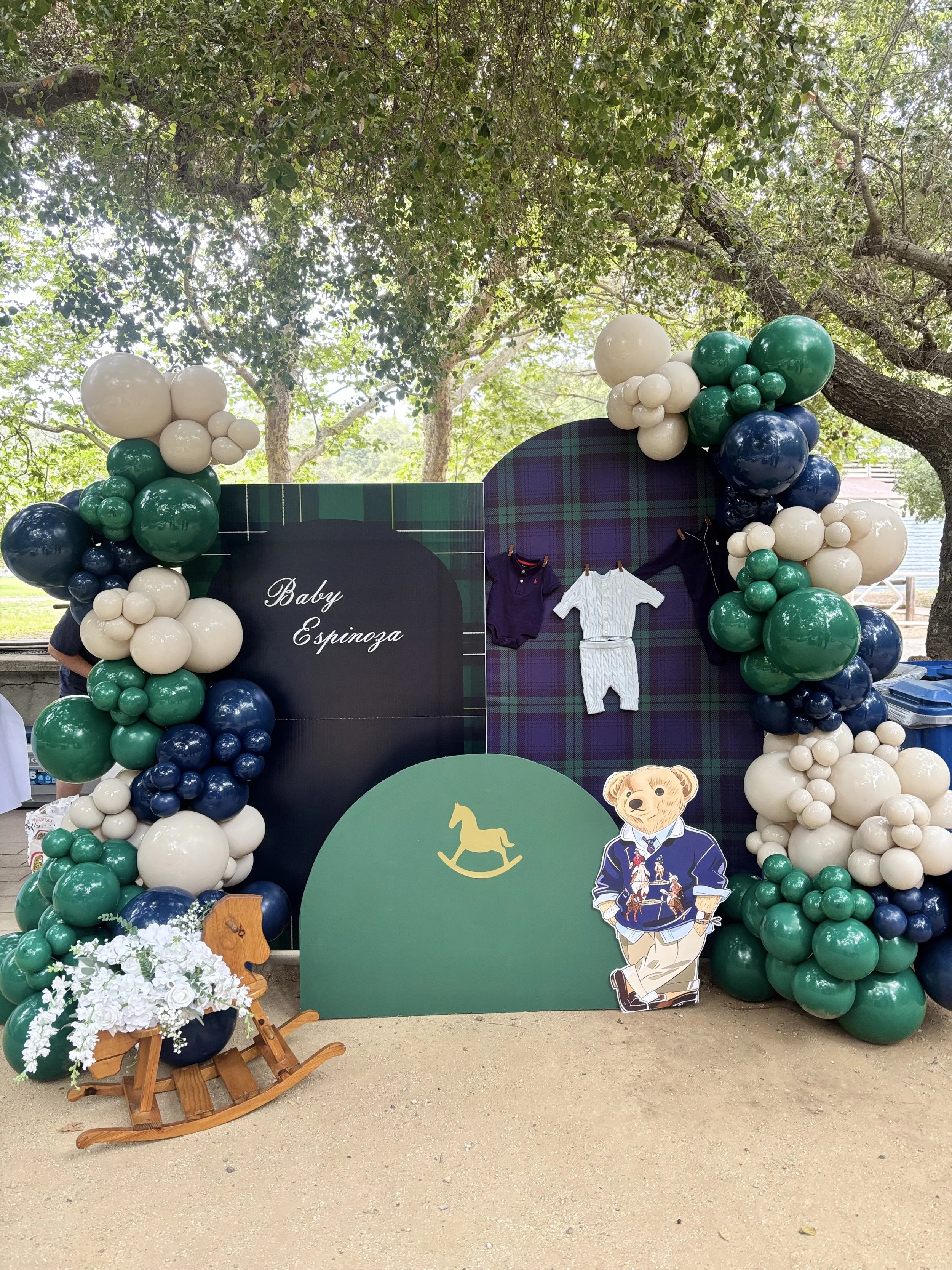 Decorative backdrop for a baby shower with green, white, and navy blue balloons, a plaid patterned arch, and a sign with the name "Baby Espinoza." There are also paper cutouts of a dog and a bear, and a small wooden rocking horse with a floral arrang