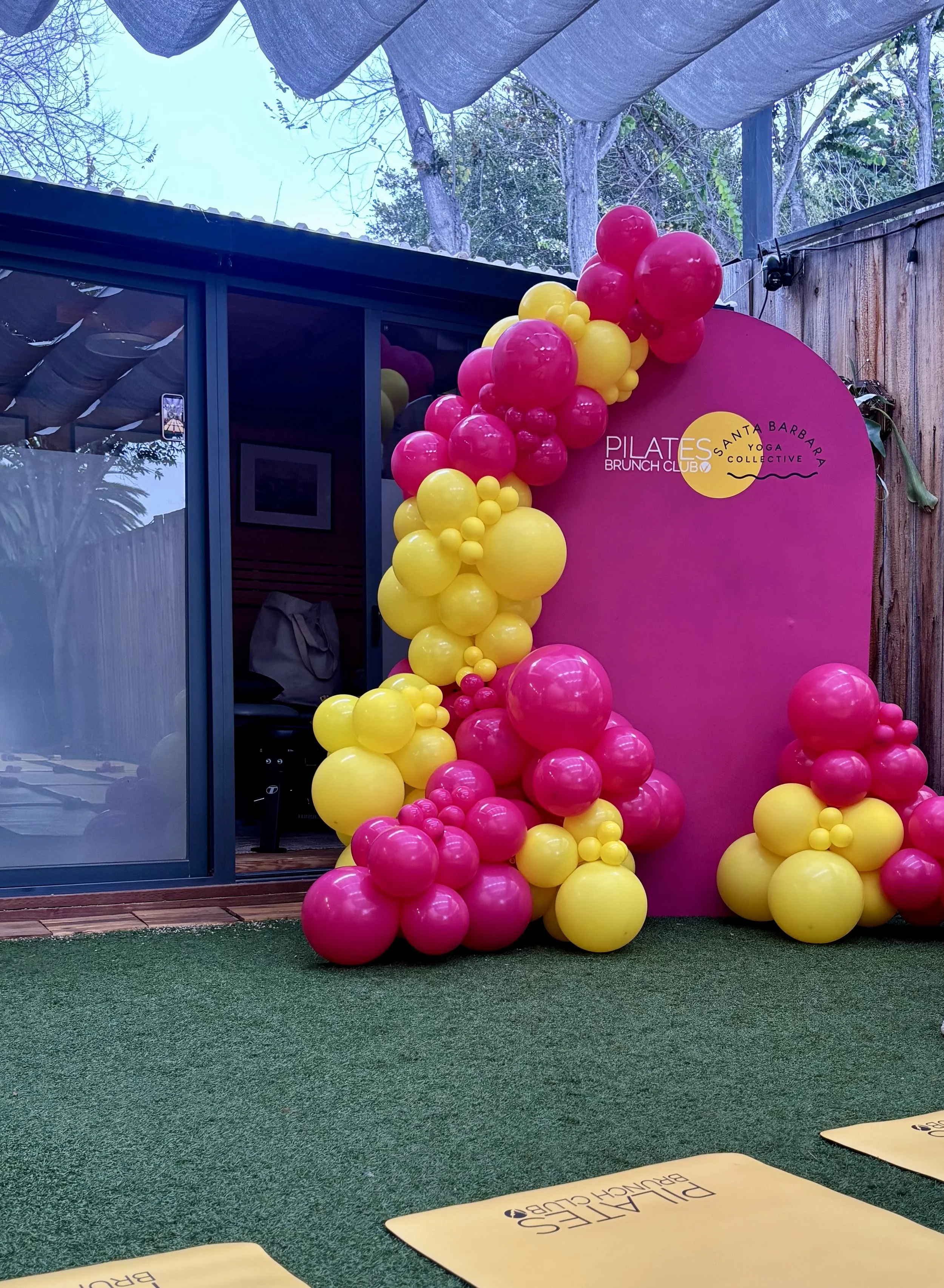 Decorative pink and yellow balloons arranged in a cluster near a pink backdrop with a Pilates studio logo, set in an outdoor space with artificial grass and wooden fencing.