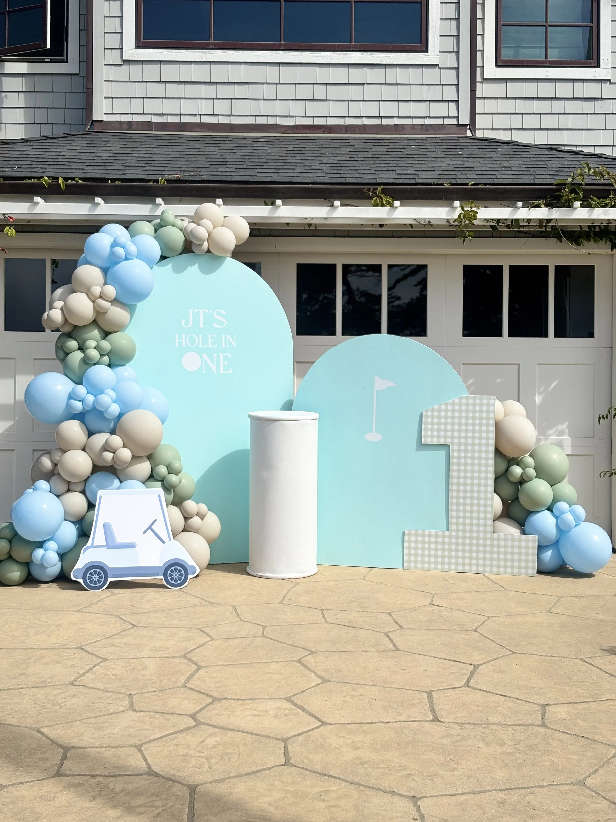 Decorative party setup with pastel blue, white, and light green balloons, a large light blue number one with a checkered pattern, a white golf cart cutout with lemon slices, and a light blue background with a small flag and cloud icon, in front of a 
