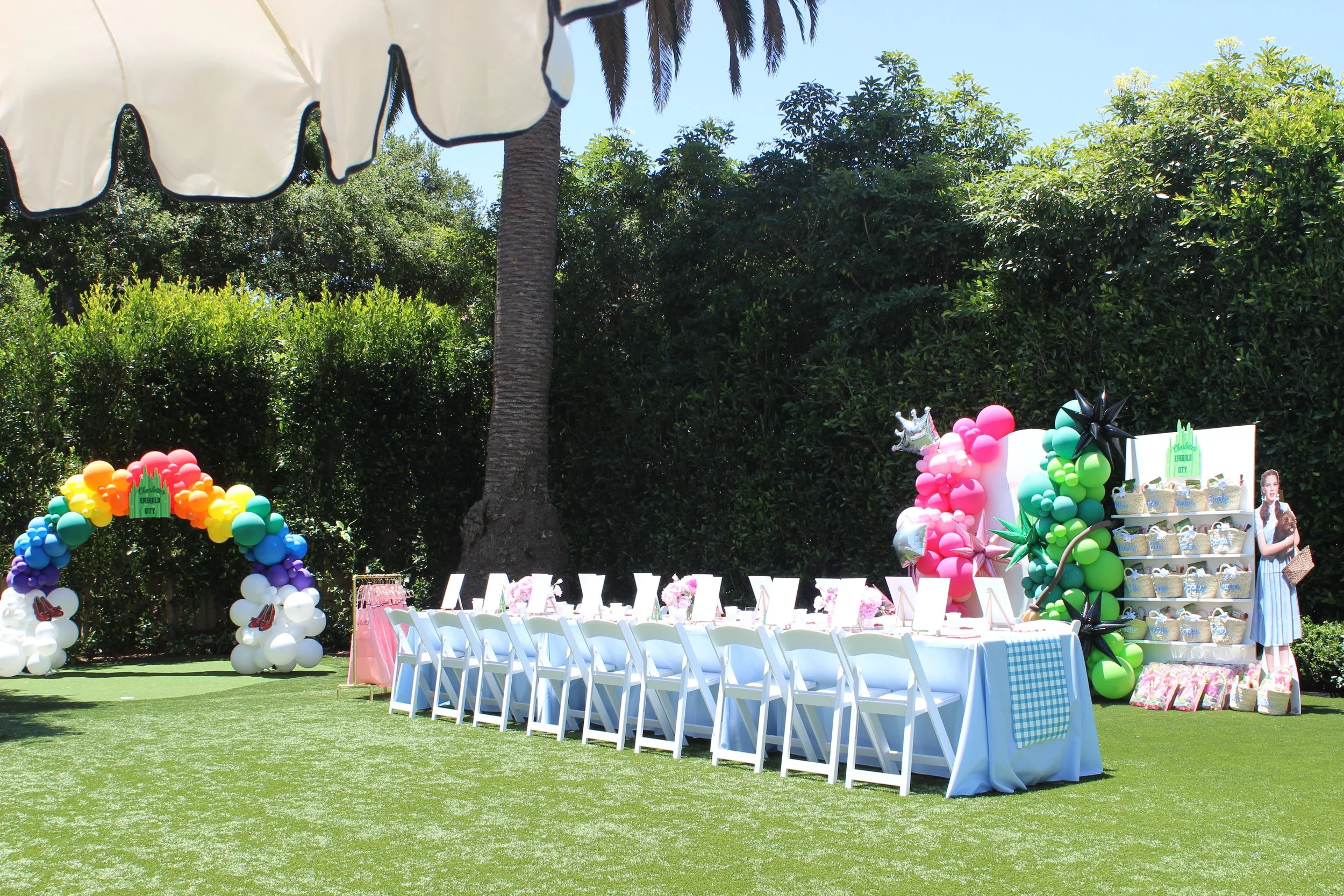 Outdoor celebration setup with a long table, decorated with colorful balloons, flowers, and themed items, under a large umbrella, surrounded by greenery and tall trees.