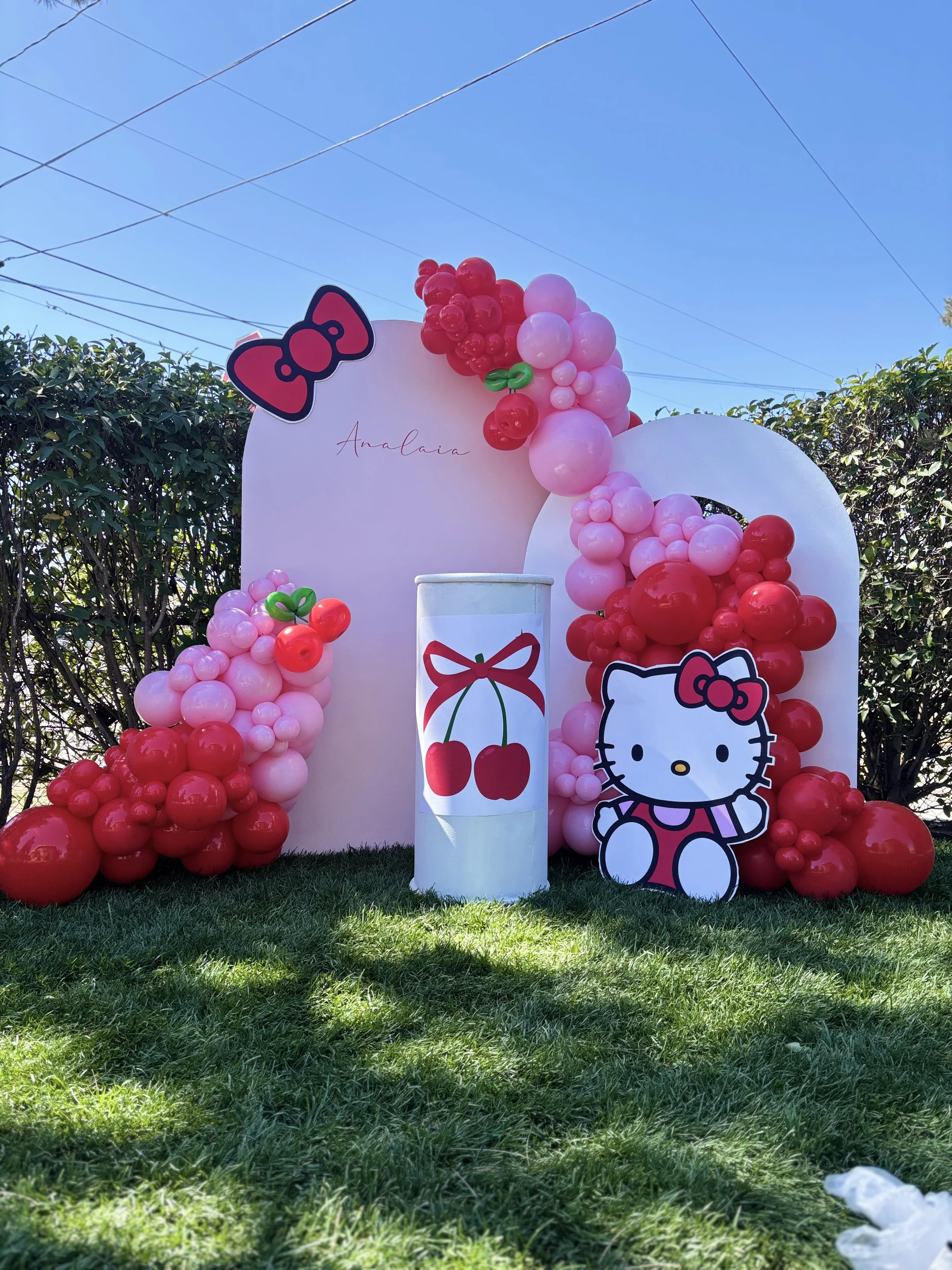 A decorative outdoor setup with pink and red balloons arranged around a white backdrop with the name 'Analaia' and a Hello Kitty cutout. The backdrop features a graphic of cherries with a red bow, and there are green balloon accents resembling leaves