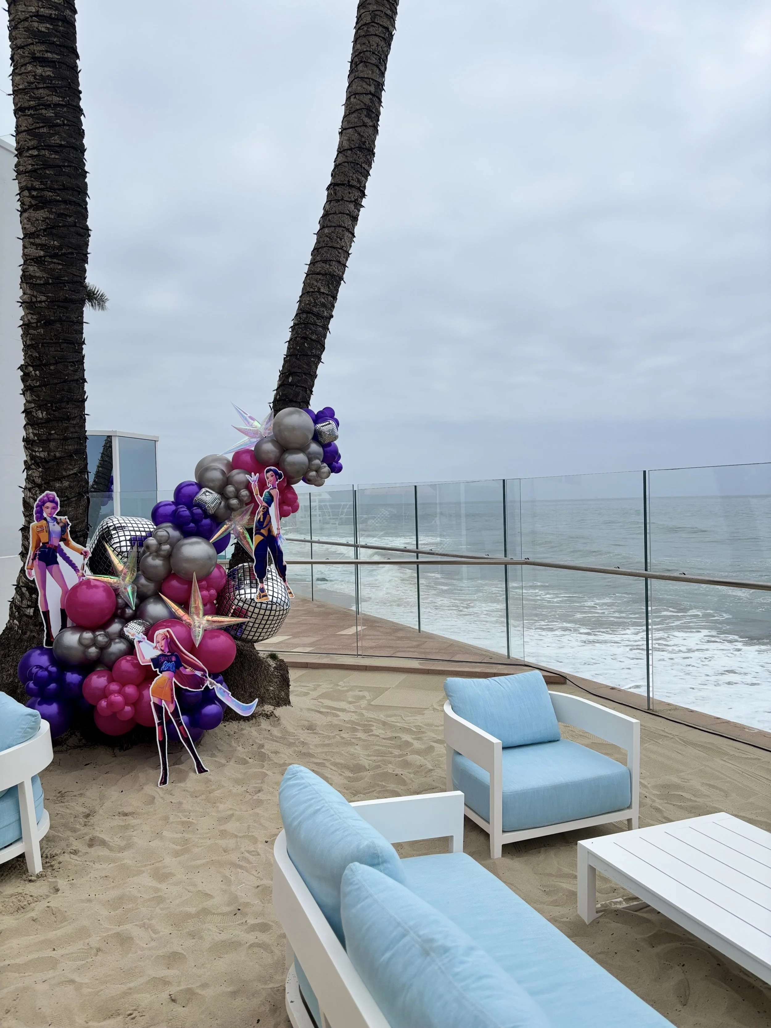 Beachside seating area with white chairs and blue cushions, decorated with colorful balloons and paper cutouts of characters, near the ocean with palm trees and cloudy sky in the background.
