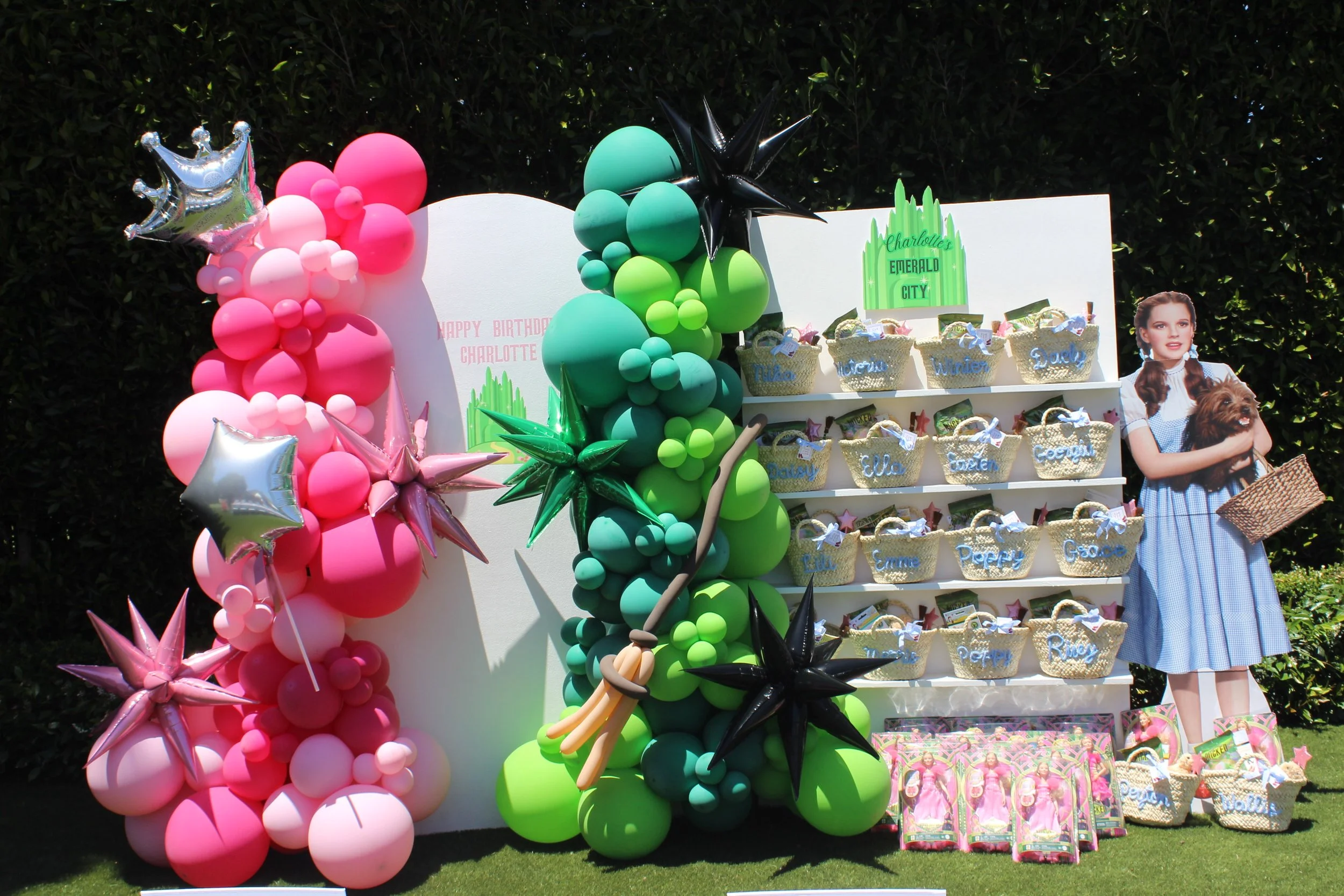Colorful balloon decorations, including pink, green, and black balloons, surrounding a white board with birthday messages and pinned baskets of small gifts with stuffed animals, a cutout of a girl holding a dog, and an assortment of party favors on a