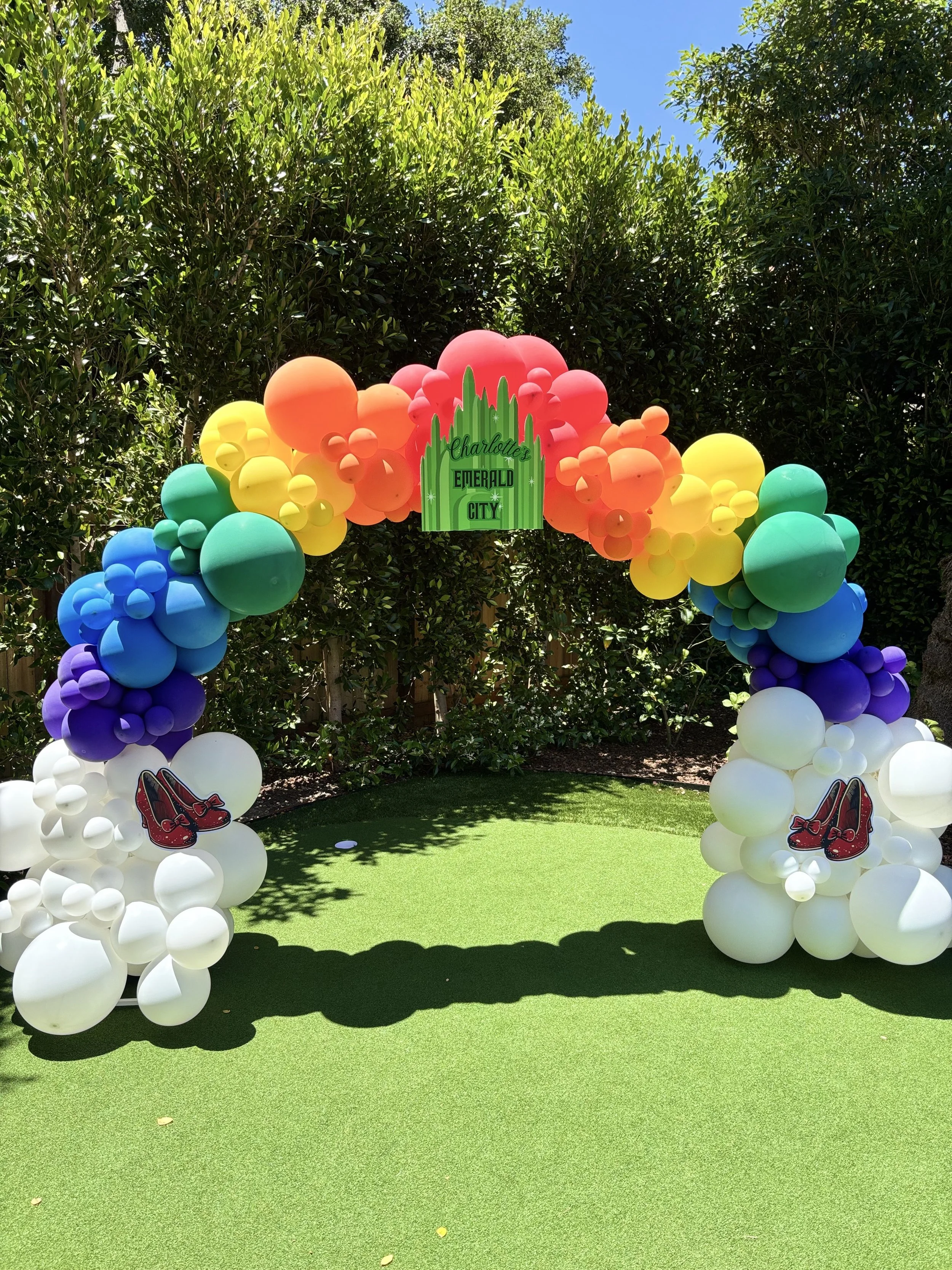 Colorful balloon arch with a sign reading 'Charlotte's Emerald City' in a backyard with green artificial turf and trees.