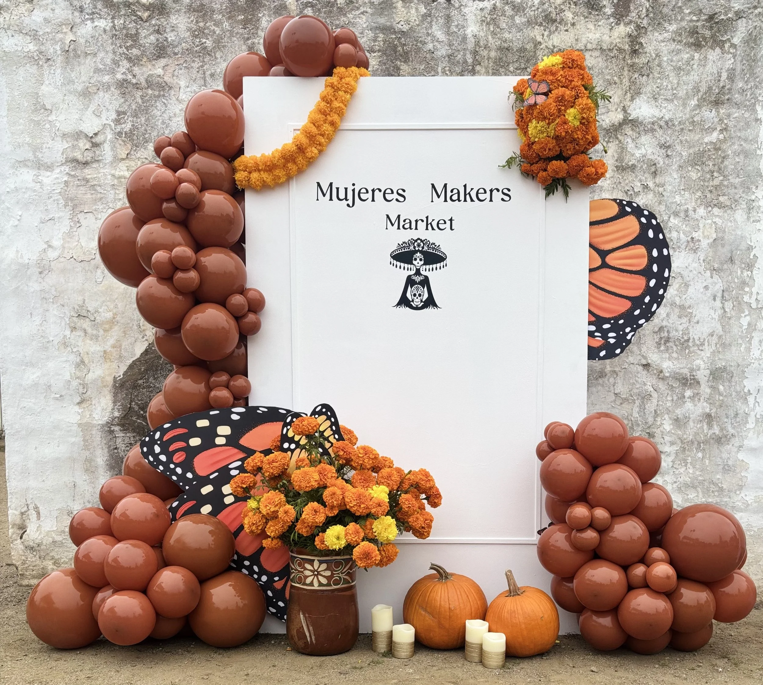 An altar decorated with orange marigold flowers, balloons, pumpkins, a sign for Mujeres Makers Market, and butterfly decorations.