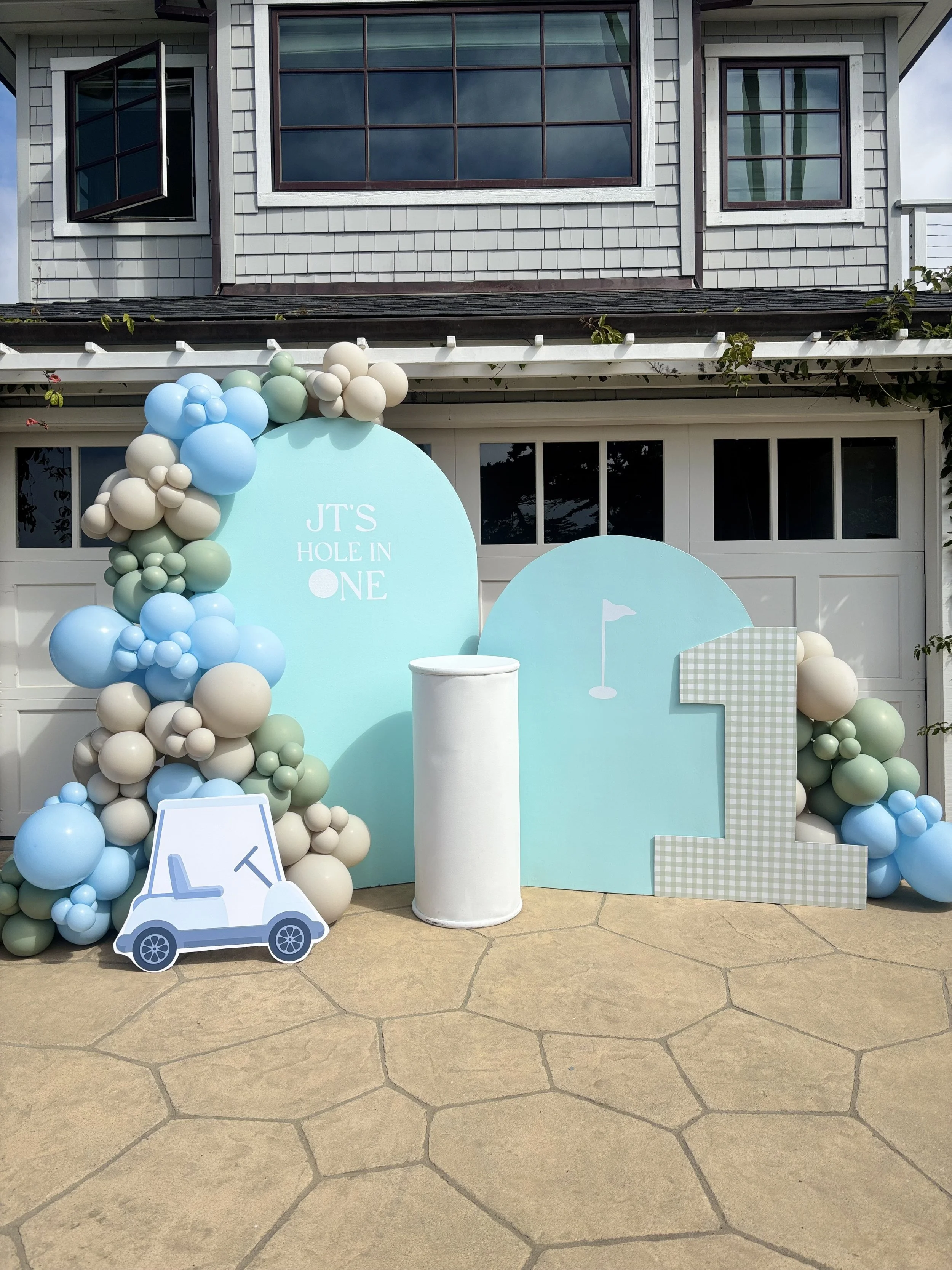 Decorative setup with balloons, a circular sign saying "Jt's Hole in One"," a small golf cart cutout, and a large number "1" sign, likely for a golf-themed celebration outside a house.