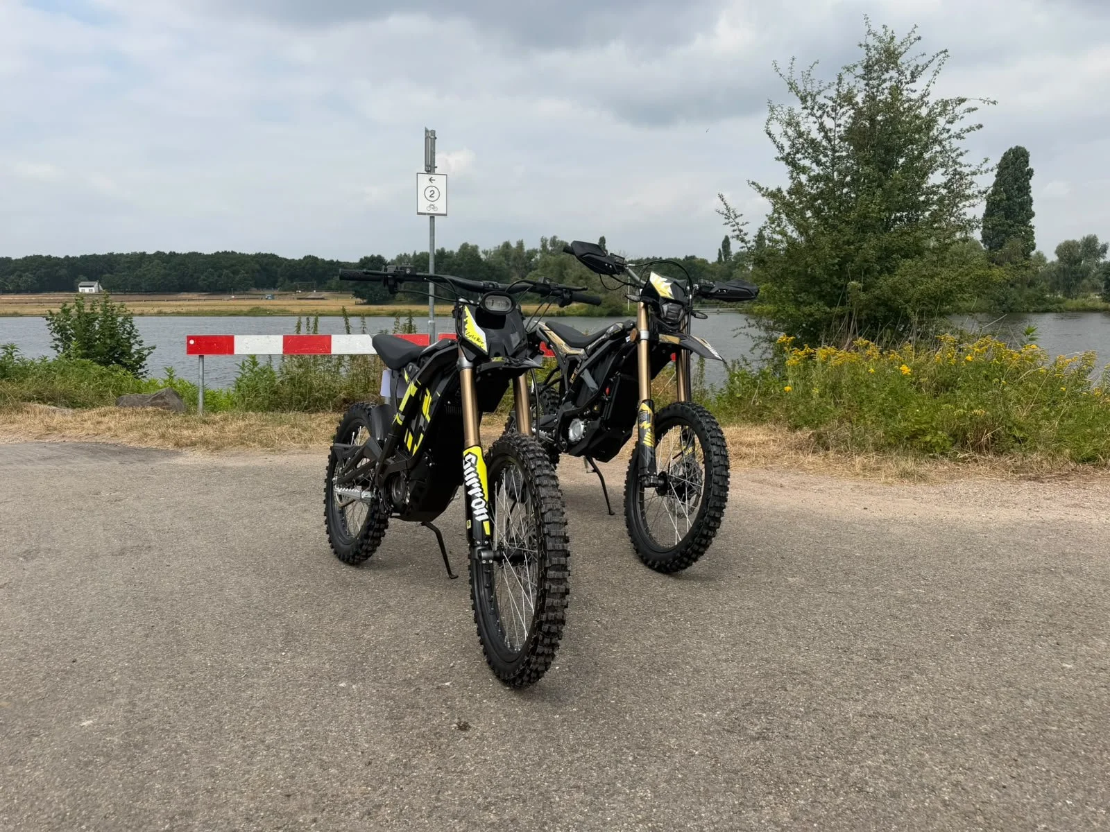 Twee elektrische mountainbikes geparkeerd aan de rand van een weg, met water en bomen op de achtergrond.