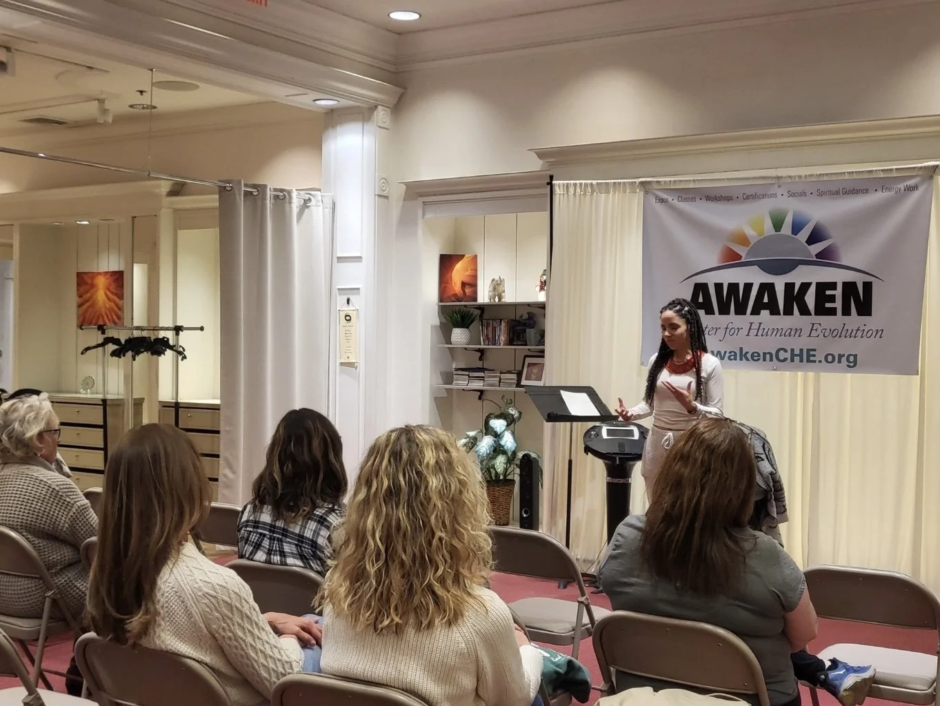 A woman giving a presentation based on energy in a room with an audience seated in chairs. There is a banner that reads 'AWAKEN' behind her and various decorative items and shelves nearby.