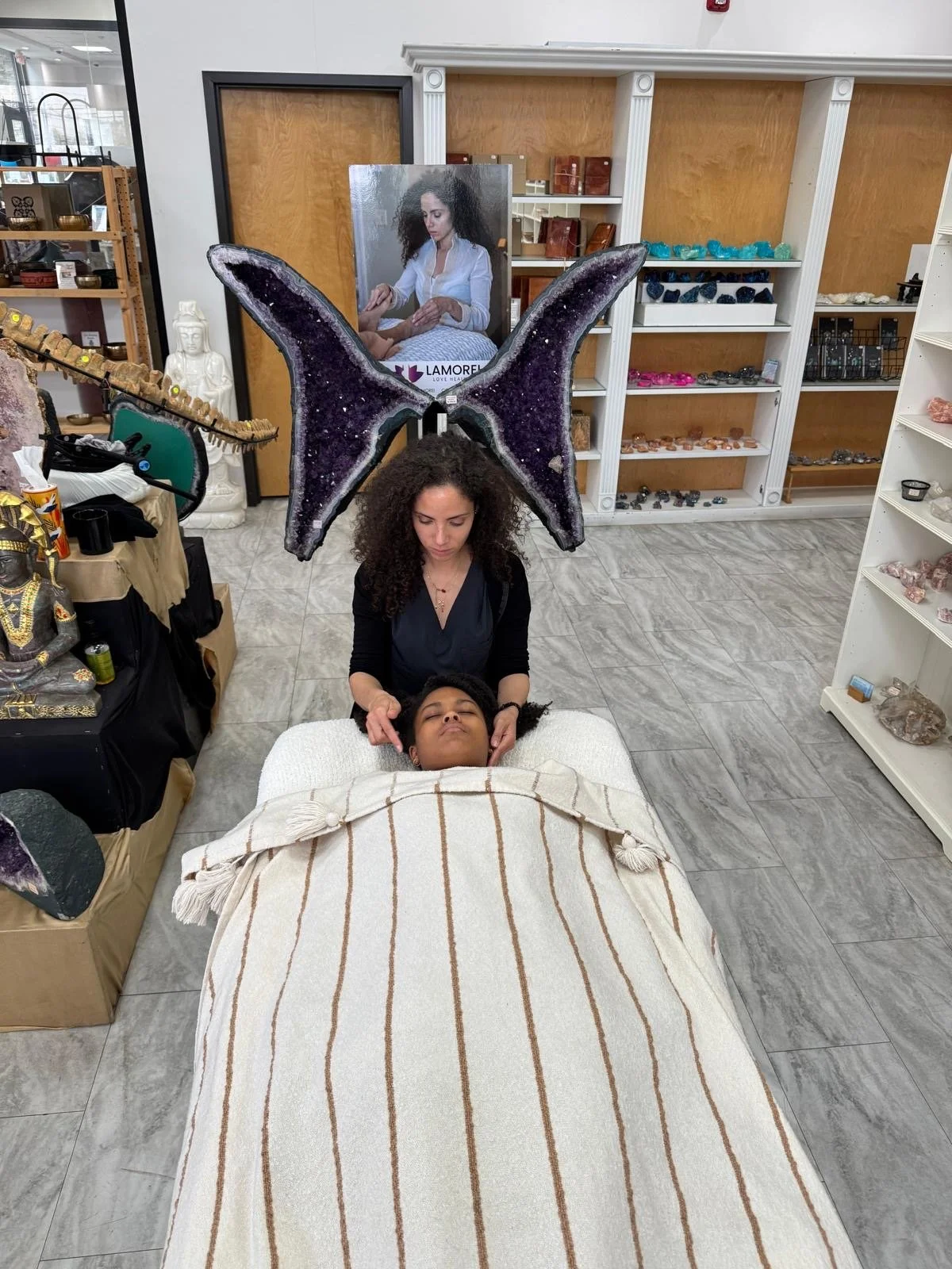 A woman with large purple gemstone wings is giving a massage  and energy therapy to another woman lying on a bed covered with a white and beige striped blanket. The setting appears to be a store or a studio with various crystals and decor around.