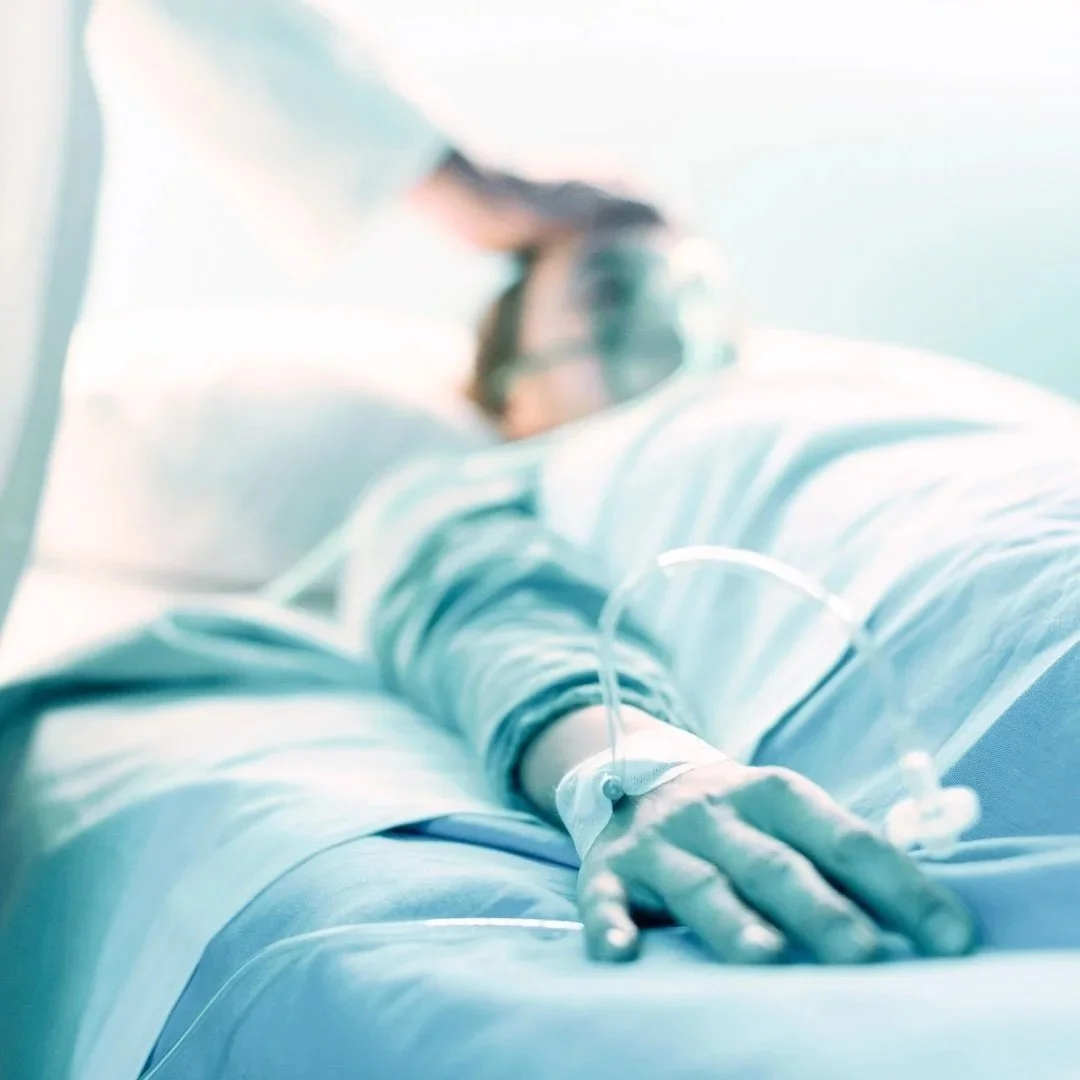 Close-up of a patient's hand with medical tubing attached, lying on a hospital bed, with a blurred background of medical  and reiki staff.