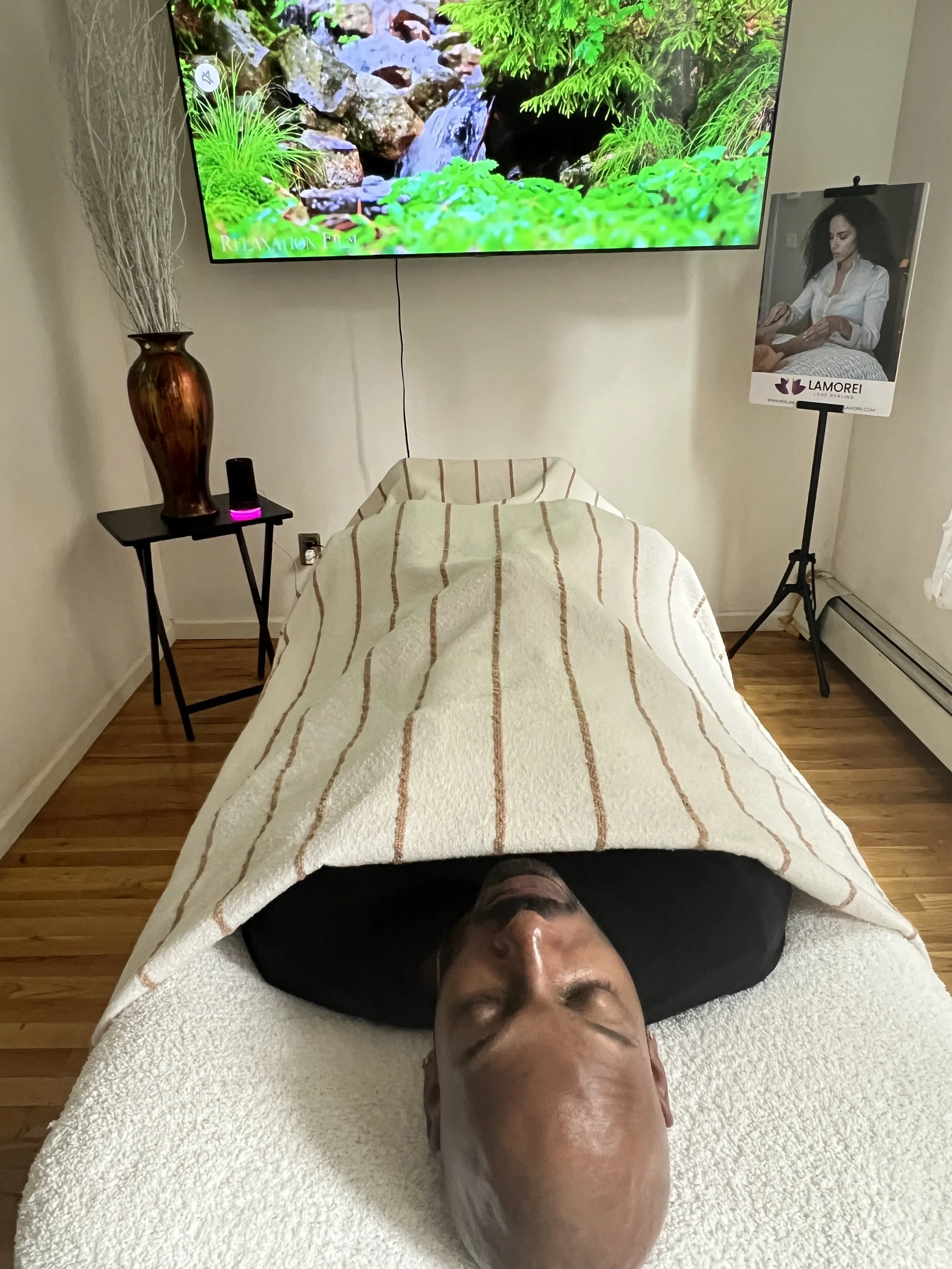 A person lying on a massage table with a white towel, receiving a massage in a room with a wall-mounted TV displaying a nature scene, a small black table with a vase and a purple diffuser, and a stand with a promotional poster for Lamorei skincare products.