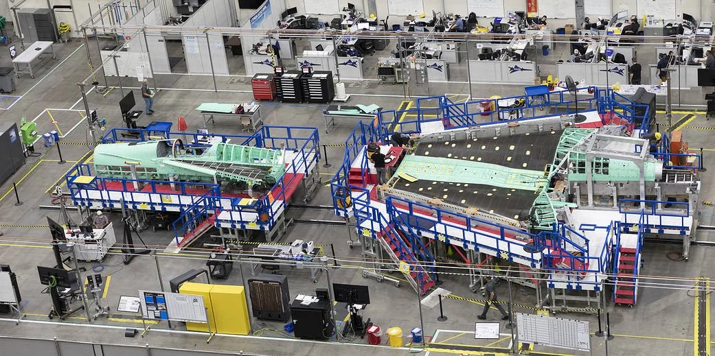 NASA's X-59 at assembly at Lockheed Martin Skunkworks in Palmdale, CA with workers and equipment, including sections of the aircraft fuselage on specialized platforms, surrounded by safety barriers.