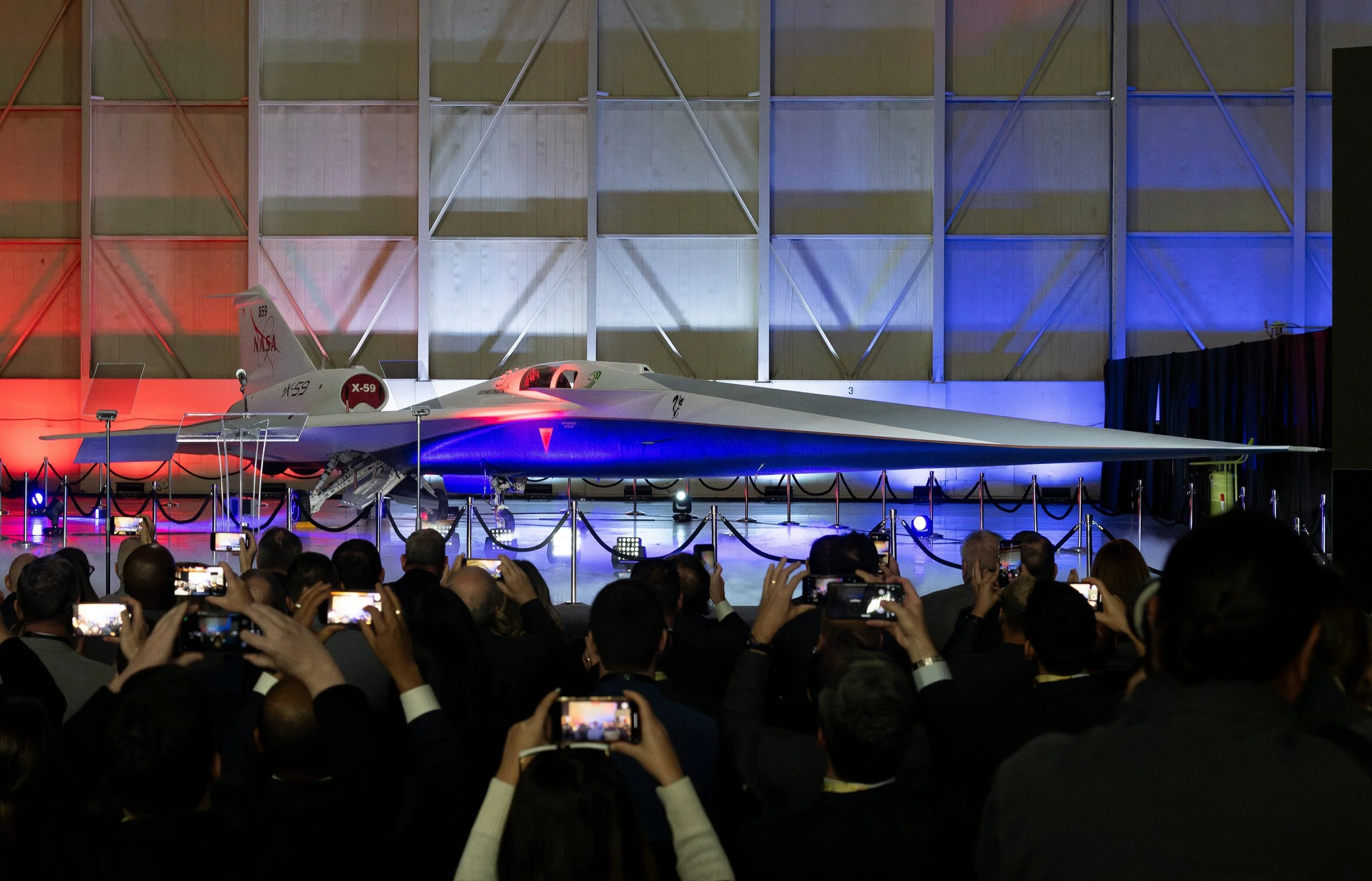 A crowd of people taking photos or videos of a sleek, futuristic aircraft (NASA X-59) on display at rollout event, with the air vehicle illuminated by colored lights.