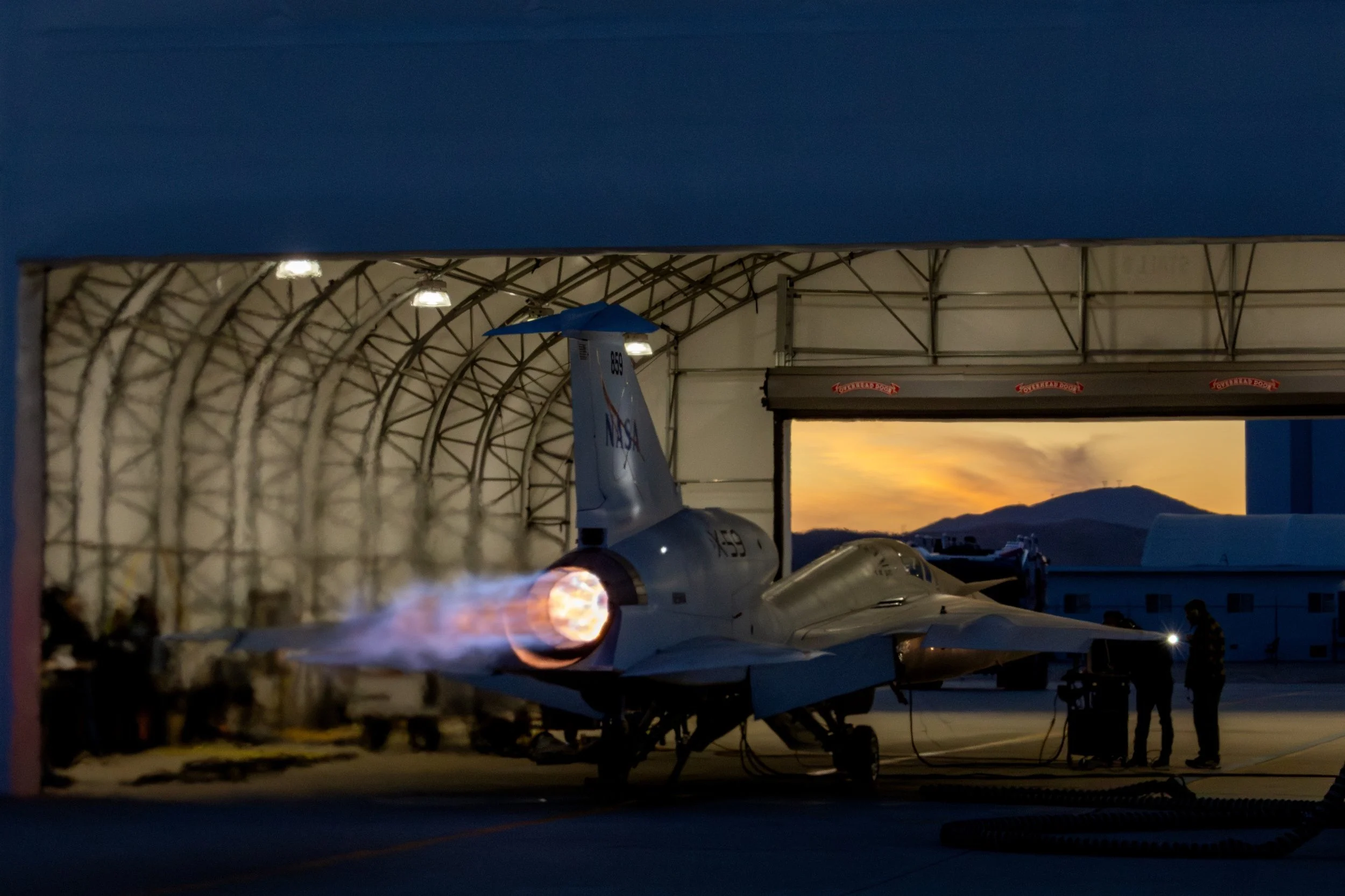 NASA X-59 aircraft on engine test inside a hangar with a sunset view outside, and two people observing the aircraft.