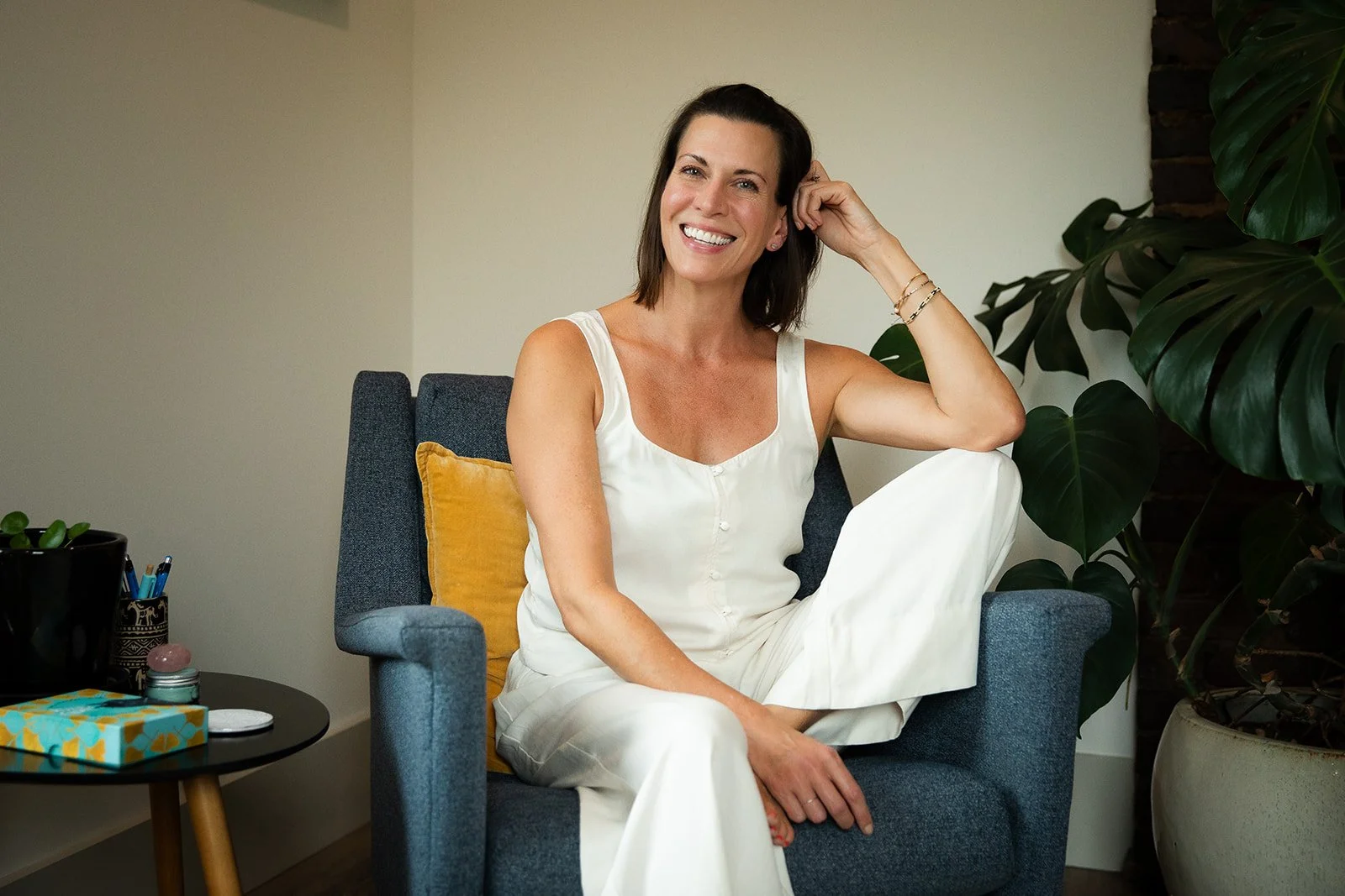 A woman with shoulder-length dark hair smiling and sitting in a gray armchair in a well-lit room, wearing a white tank top and white pants. There are large green plants on the right and a small round table with office supplies and a gift box on the left.