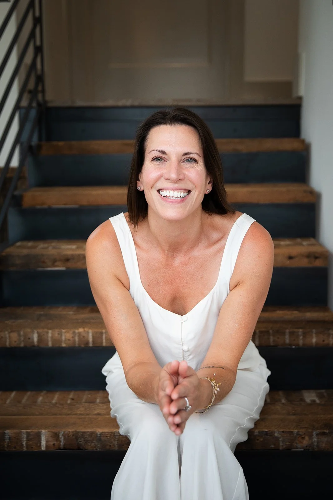 Asheville eating disorder therapist Caitlin Craig sitting on stairs, smiling with her hands clasped in front of her, wearing a white sleeveless top and white pants.
