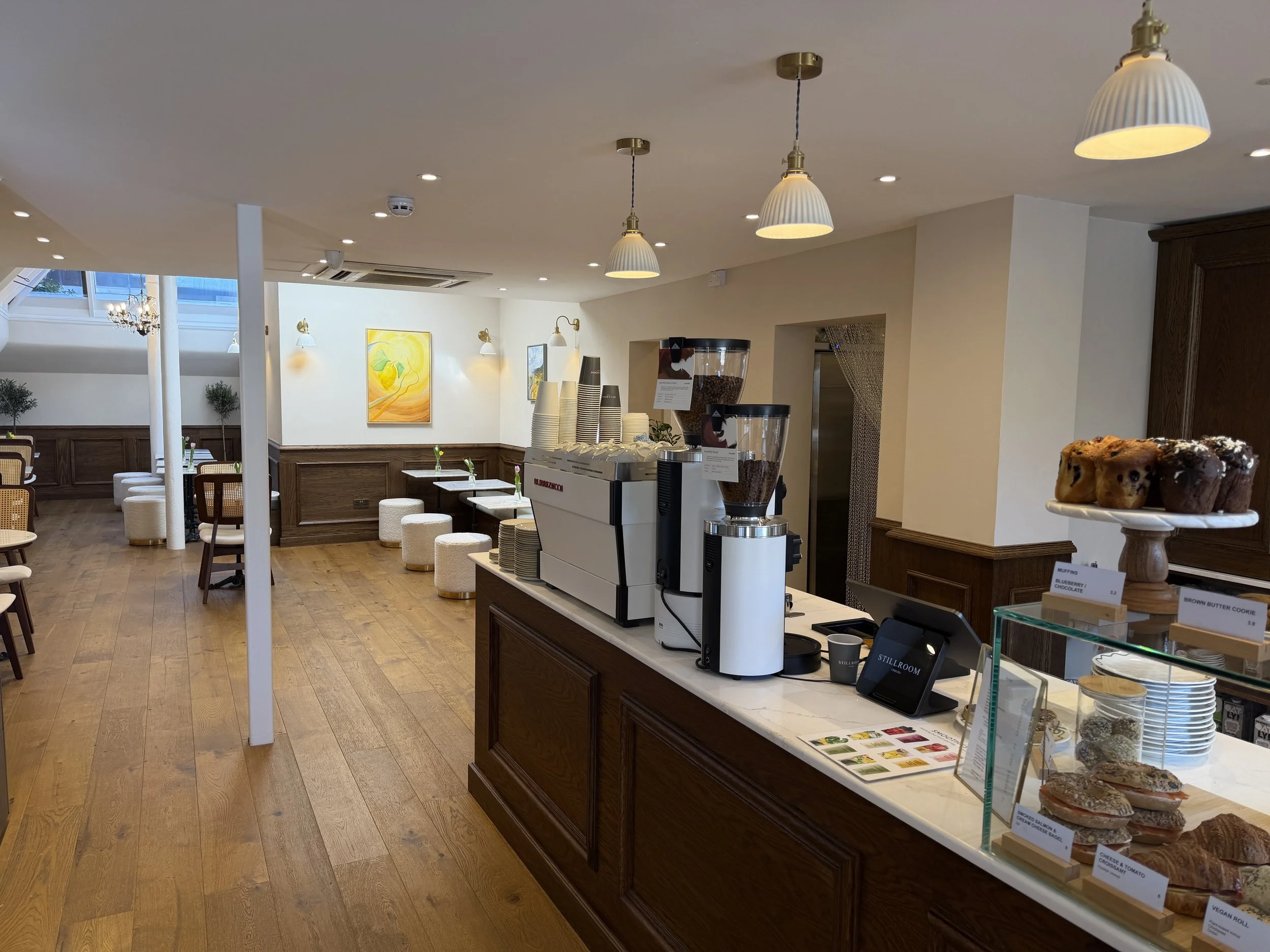 Interior of a cafe with wooden floors and white walls, decorated with abstract art paintings, warm lighting, and a breakfast counter with coffee machines, baked goods, and menu signs.