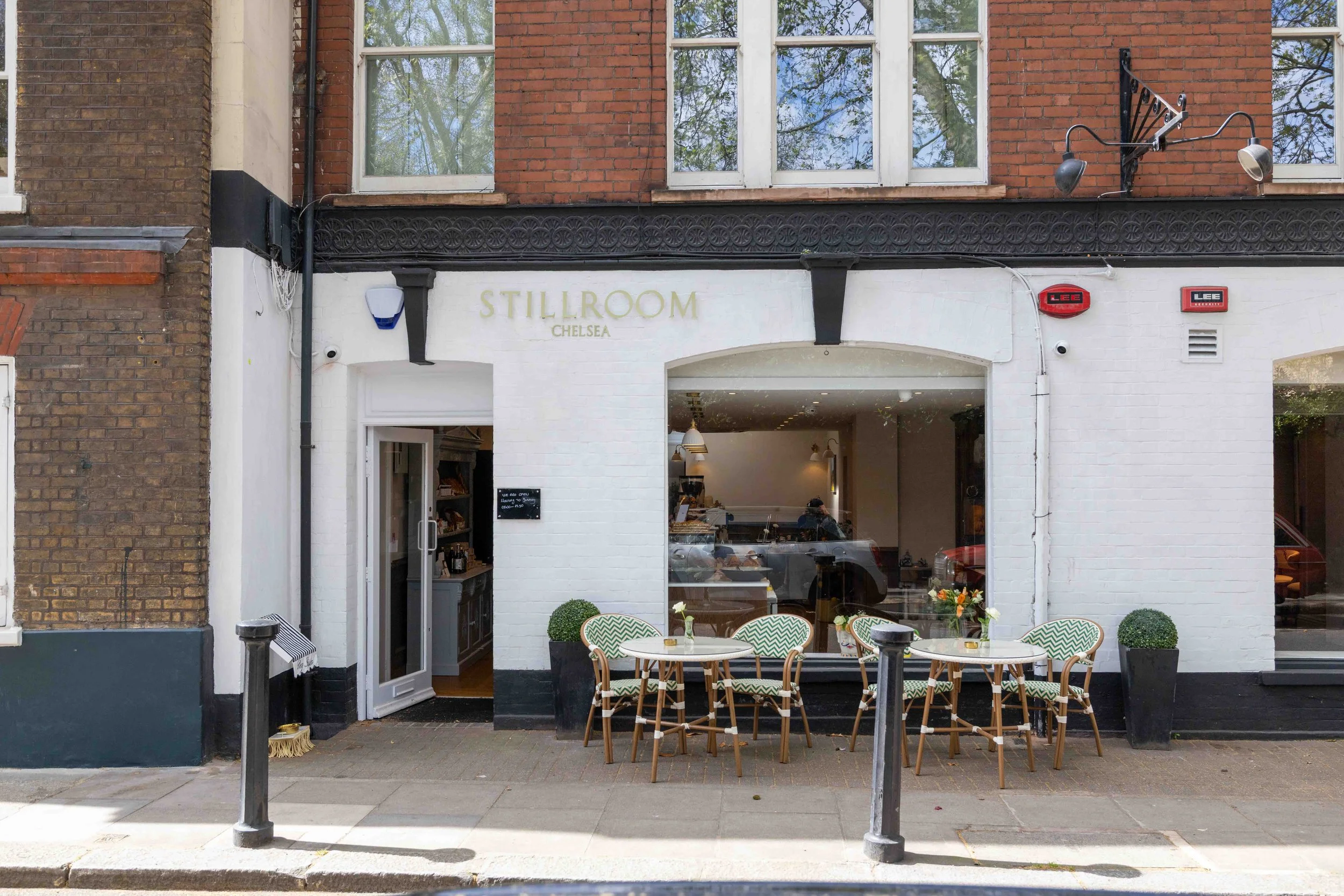 The exterior of a restaurant named Still Room Chelsea with a white brick facade and large windows. There are tables and chairs outside, set with small vases of flowers, and the interior is visible through the open door. The street in front features a sidewalk, a drinking fountain, and black bollards.