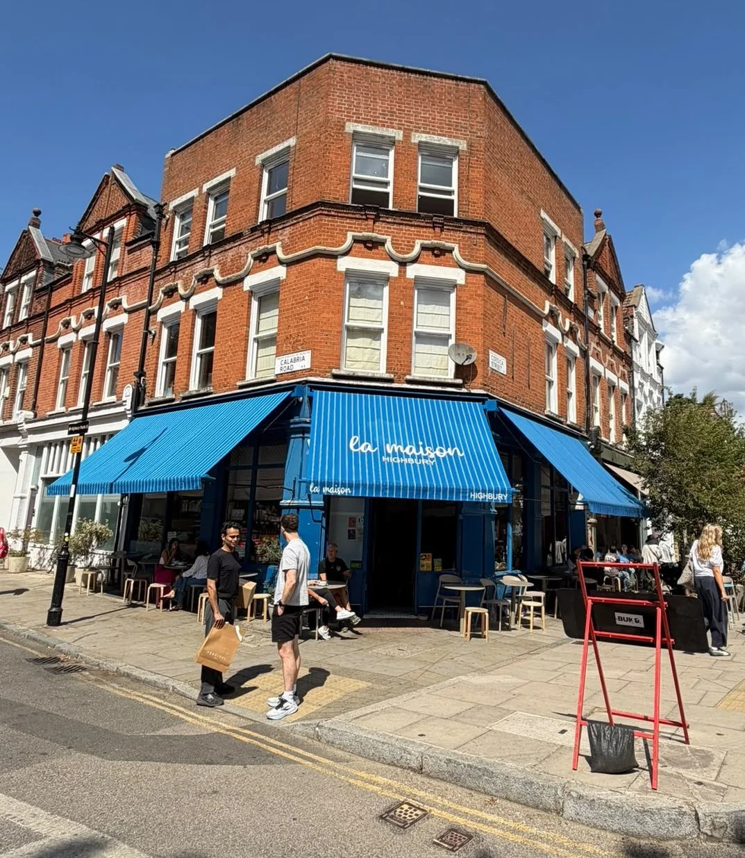 Corner storefront with a blue and white striped awning that reads 'la maison HIGHBURY,' in a red-brick building on a sunny day with people walking and sitting outside.
