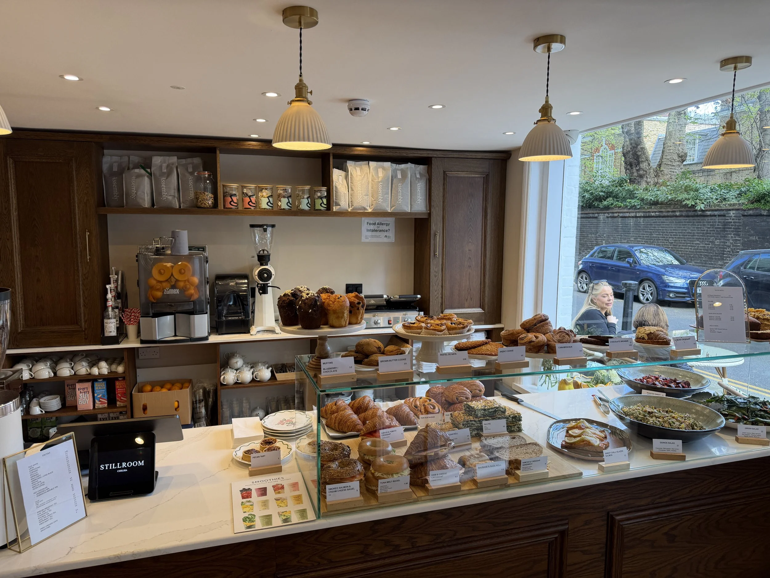 A bakery counter with assorted baked goods including muffins, croissants, and slices of bread, along with label tags and a display case of salads. There is a wooden shelf with packaged coffee and jars behind the counter, and large windows revealing parked cars and trees outside.