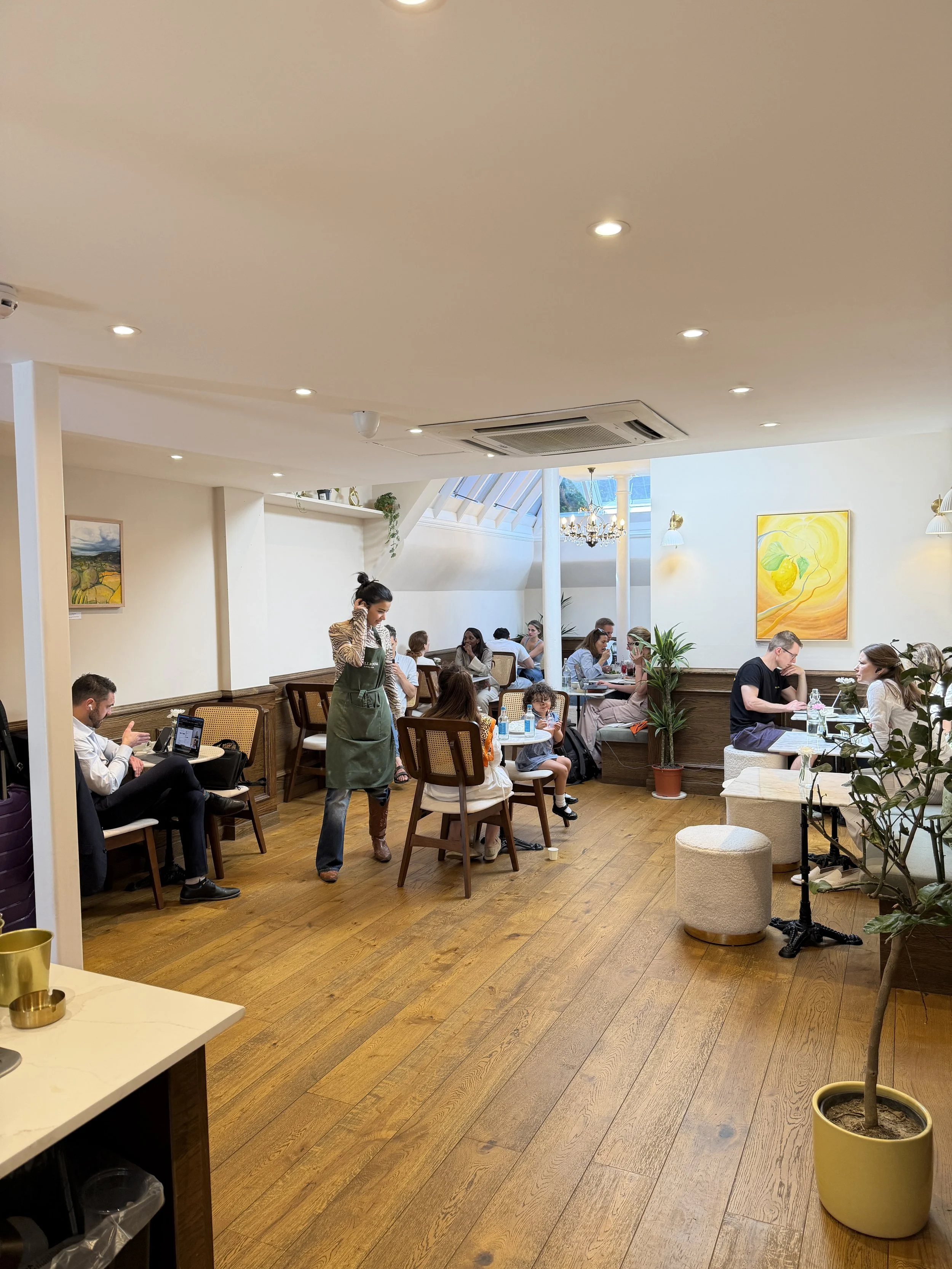 A busy Stillroom Chelsea cafe with customers sitting at tables,  and colorful David McAdam Freud paintings on the walls.
