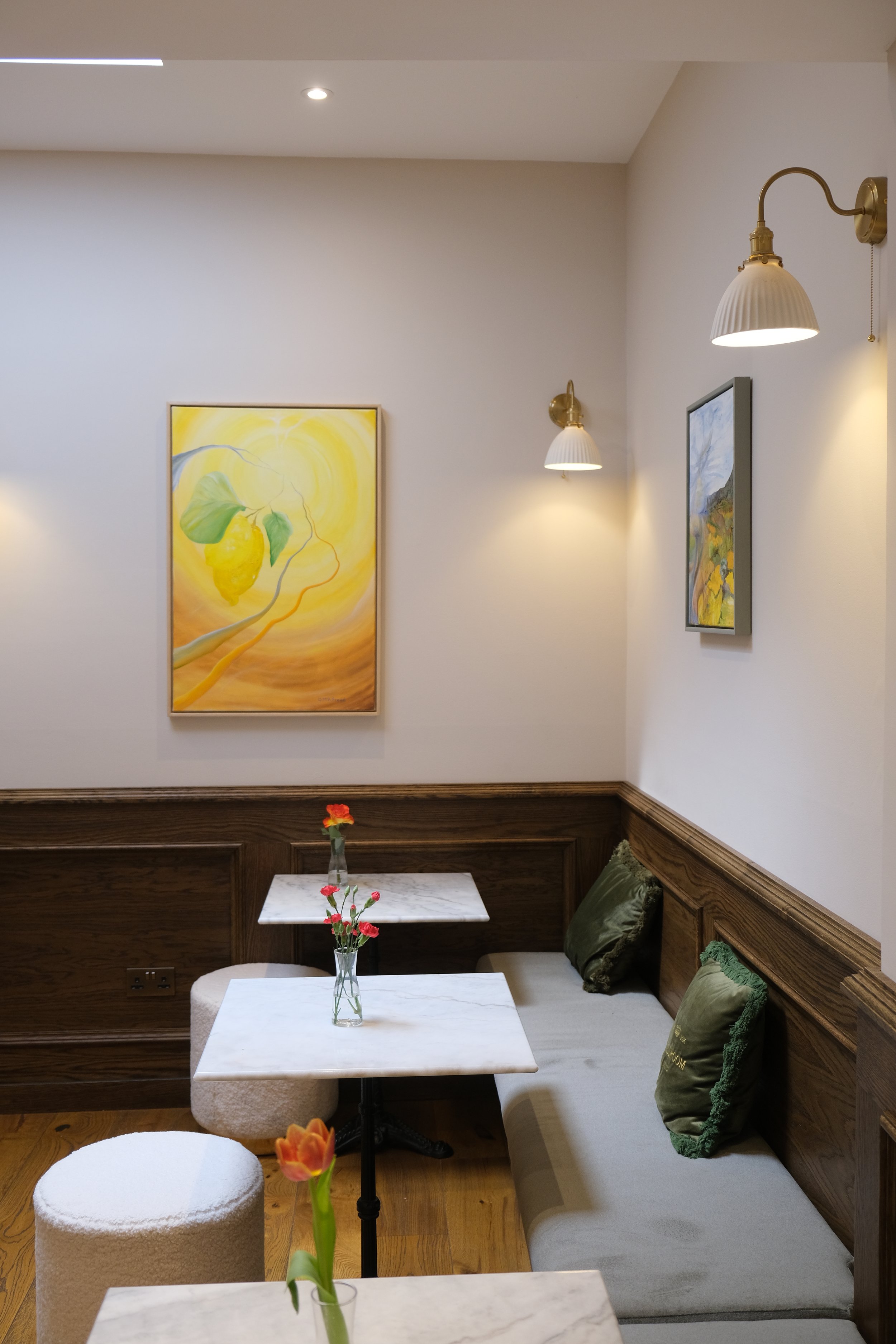 A cozy seating area in a cafe or restaurant with white marble tables, green pillows, and a wooden wall with two framed landscape paintings. There are small vases with flowers on the tables, and wall-mounted lamps provide soft lighting.