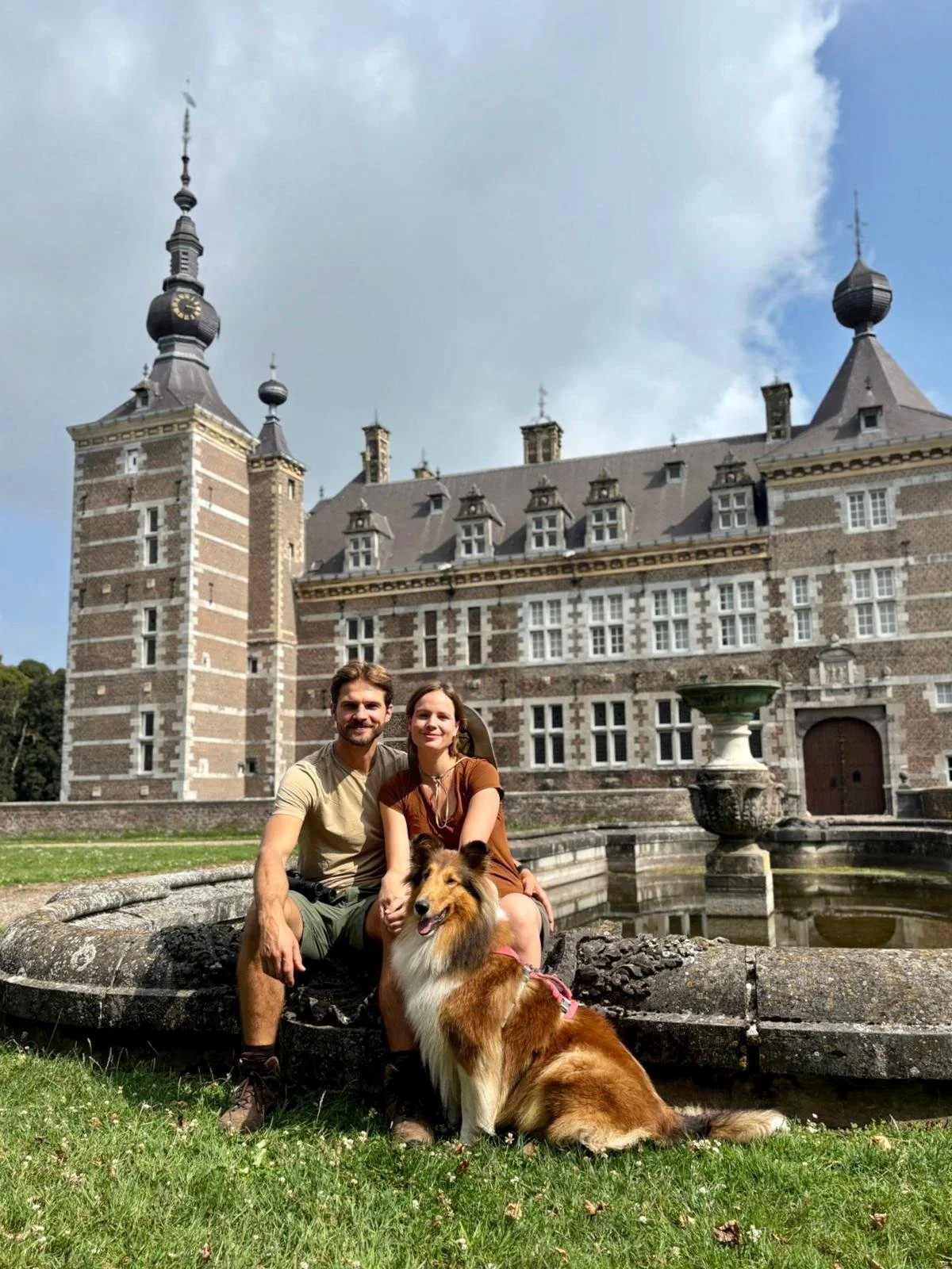 Een koppel zit voor een kasteel, samen met hun hond, in een tuin met een fontein.