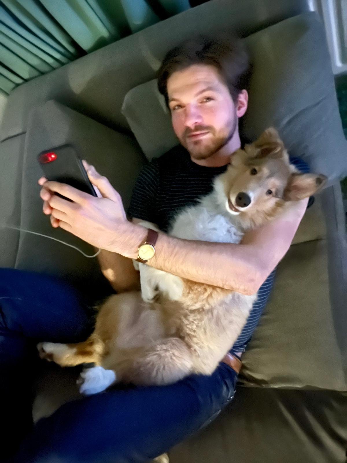 Man ligt op een bank, houdt een hond en kijkt in de camera terwijl hij een smartphone vasthoudt.
