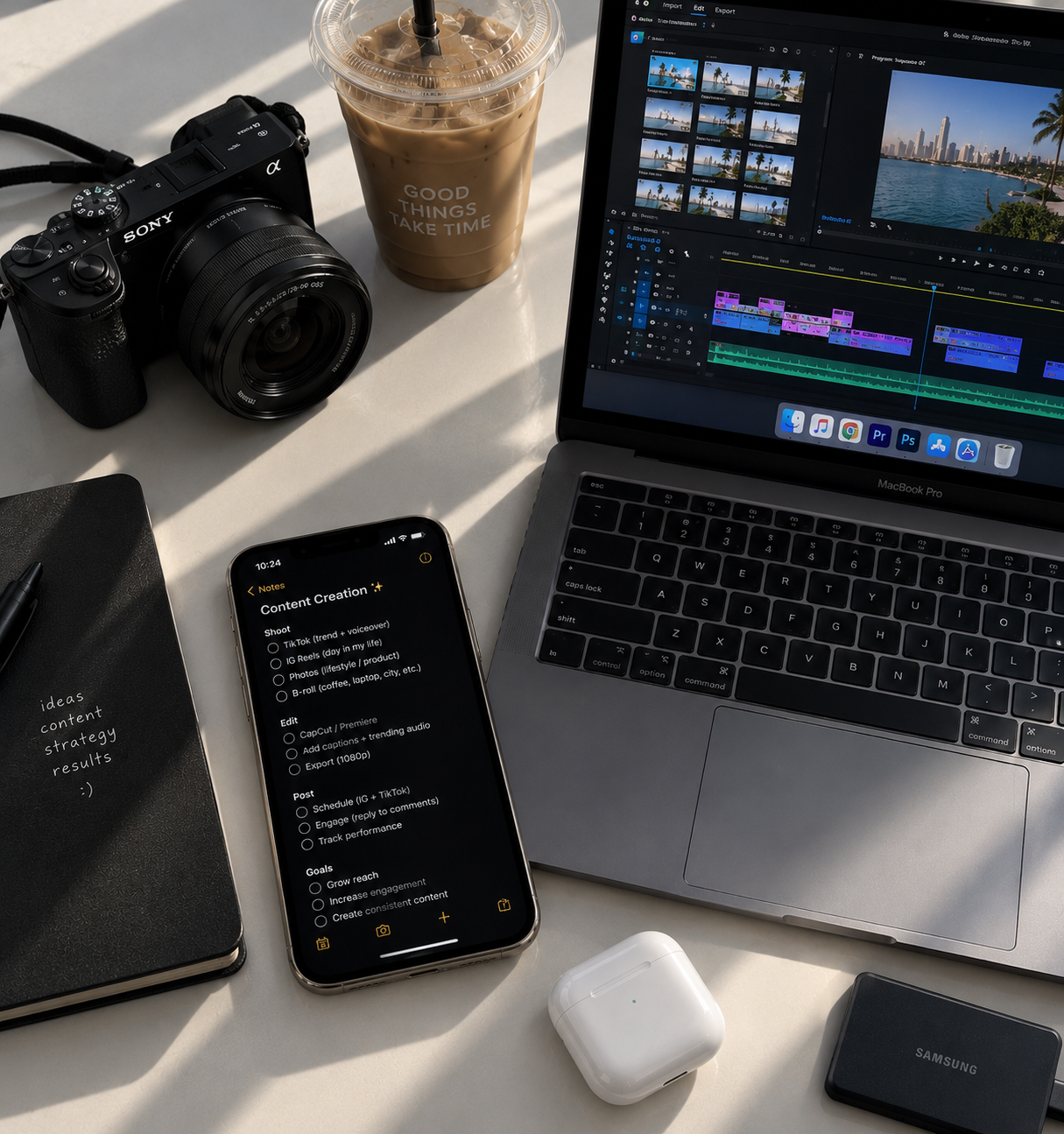 A workspace with a camera, a cup of iced coffee, a laptop displaying video editing software, a smartphone with notes, a notebook, wireless earbuds case, and a portable hard drive on a white surface.