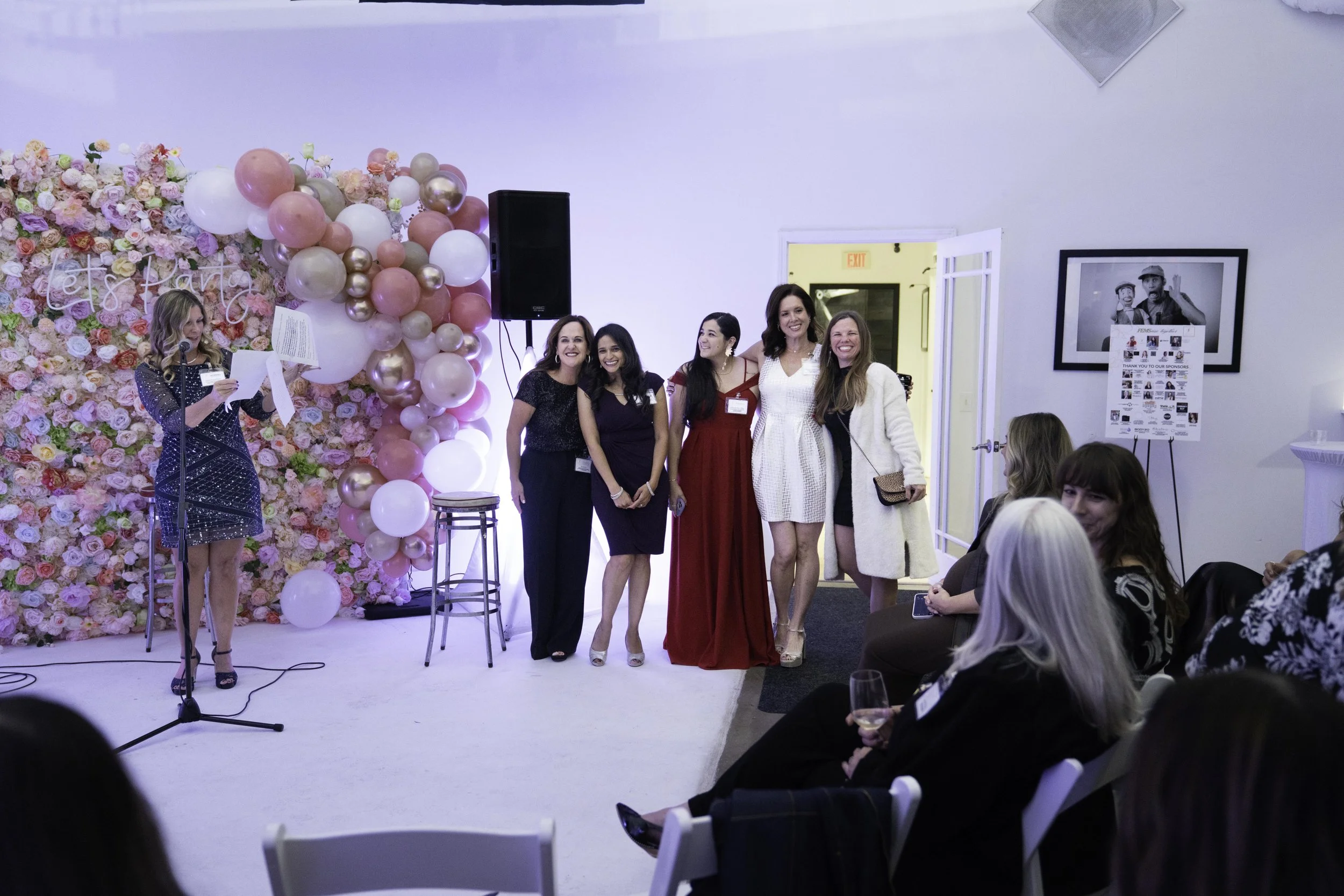 A woman in a blue dress is speaking into a microphone at a decorated event with five women posing for a photo in front of a pink and flower backdrop, with an audience seated nearby.