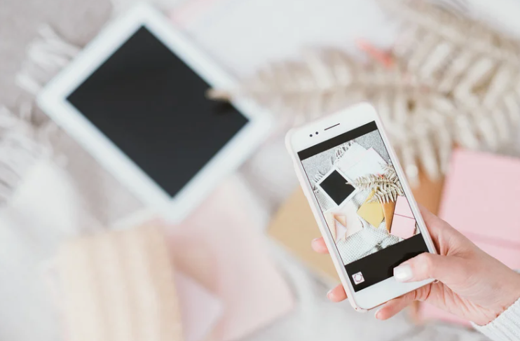 Person taking a photo of a flatlay arrangement with a smartphone, featuring a tablet, patterned fabric, and colorful paper items.