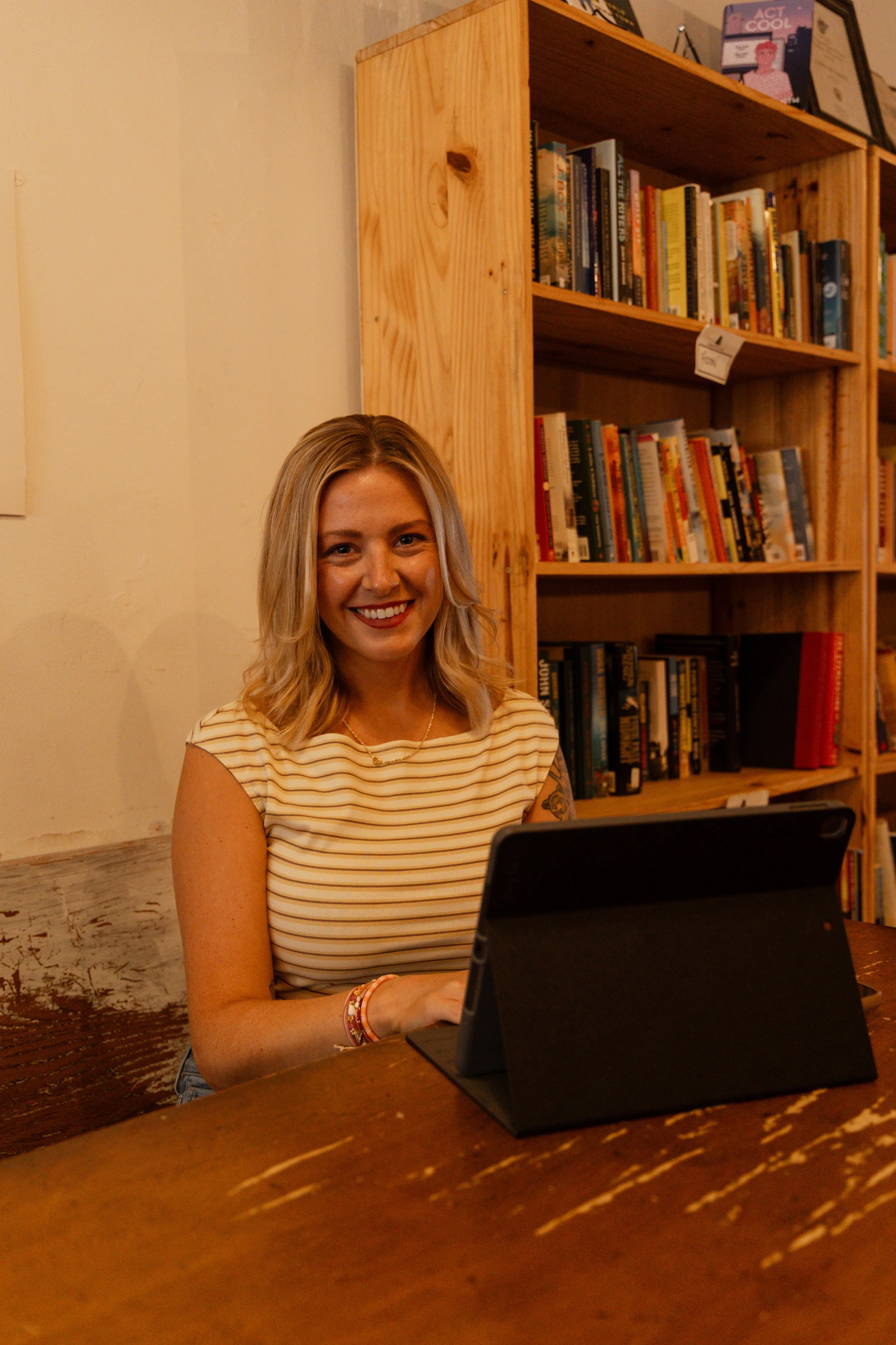 A smiling woman with blonde hair, wearing a striped yellow and white top, sitting at a wooden table with a tablet or laptop in front of her. Behind her is a wooden bookshelf filled with books, and framed art or posters on the wall.