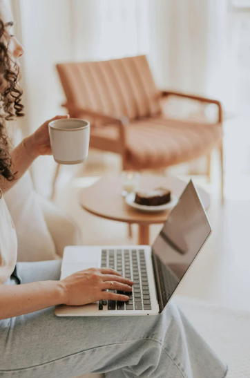 Person sitting on a couch, using a laptop, holding a coffee mug in a cozy living room with a pink sofa and coffee table.