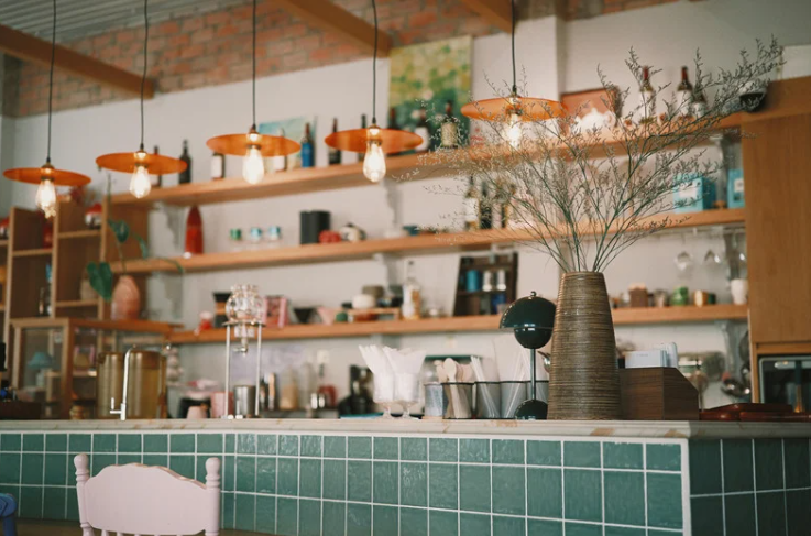 Interior view of a cafe or kitchen with hanging pendant lights, wooden shelves filled with various kitchenware and decor, and a countertop with a large vase of dried flowers.