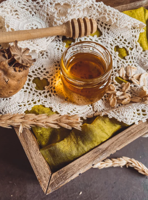 A jar of honey on a lace cloth with a honey dipper, dried wheat, and pinecones in a wooden tray