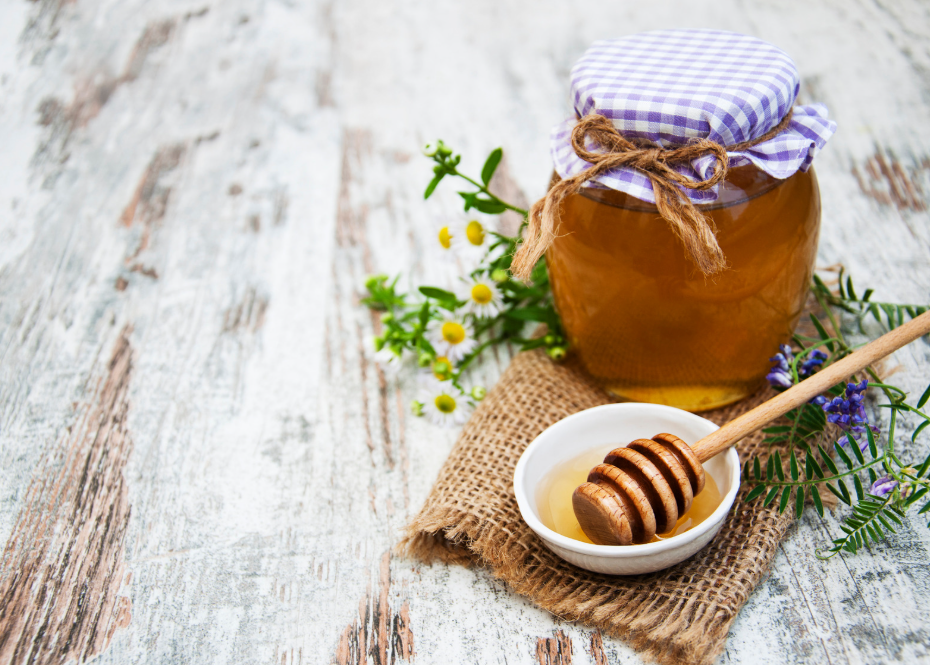 Jar of honey with a purple checkered cloth cover, a small bowl of honey with a honey dipper, and wildflowers on a wooden surface.