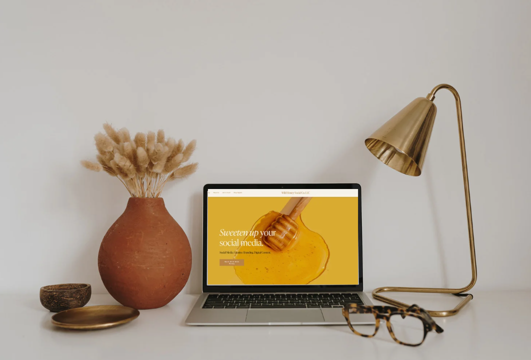 A minimalist desk setup with a laptop displaying a website about social media marketing, a gold desk lamp, a pair of glasses, a large terracotta vase with dried flowers, a small dish, and a decorative bowl, all against a white wall.