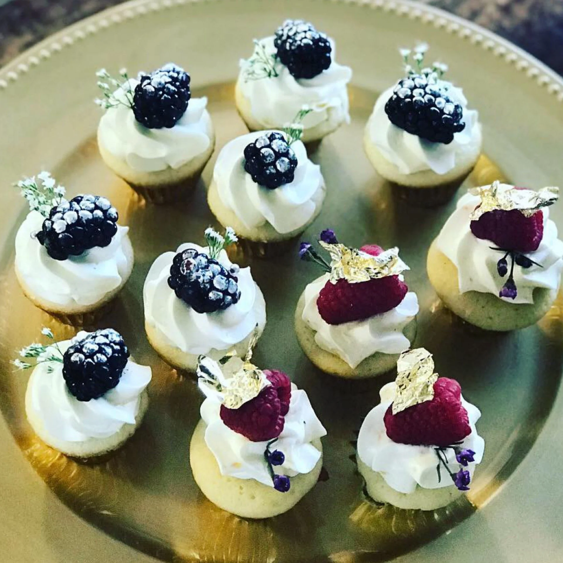 A plate of mini cupcakes topped with blackberries, and decorated with edible gold and edible flowers.