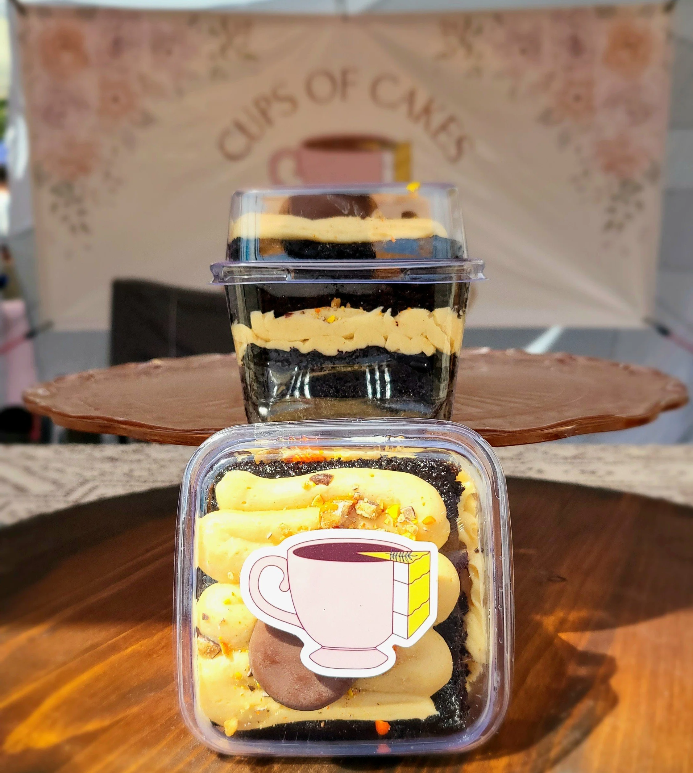 Cake Cups in a small clear plastic container. A layered cake dessert, including yellow, white, and dark chocolate layers, topped with sprinkles and nuts. A sticker with CU\ups of Cakes logo is on the front of the container.