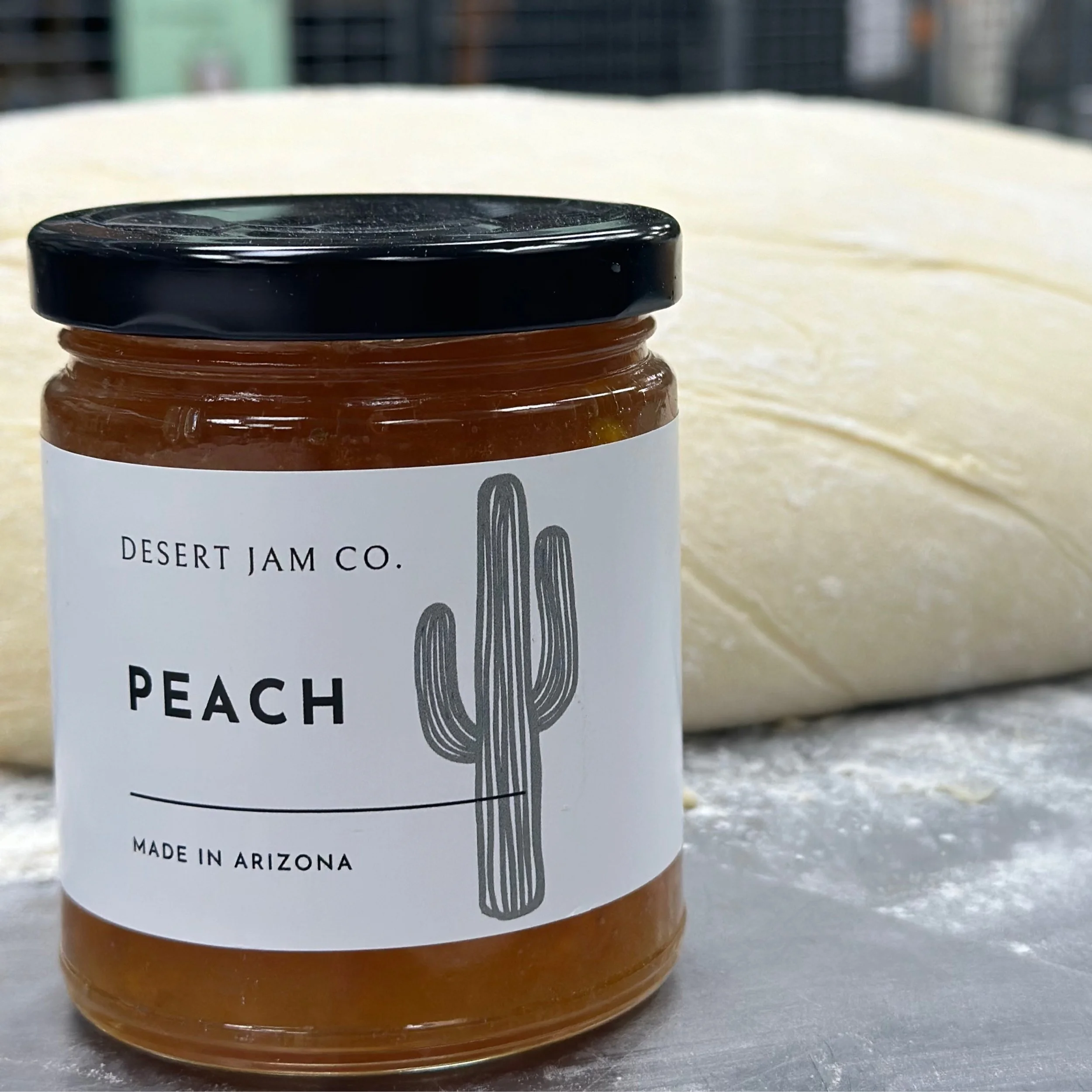 A jar of peach preserves from Desert Jam Co. on a gray surface, with a black lid and a white label featuring a black cactus illustration. The jar contains peach jam and is made in Arizona.
